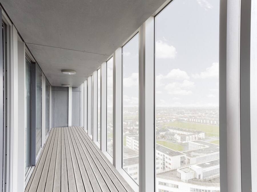 A corridor with large windows and a wooden floor, leading to a city view.