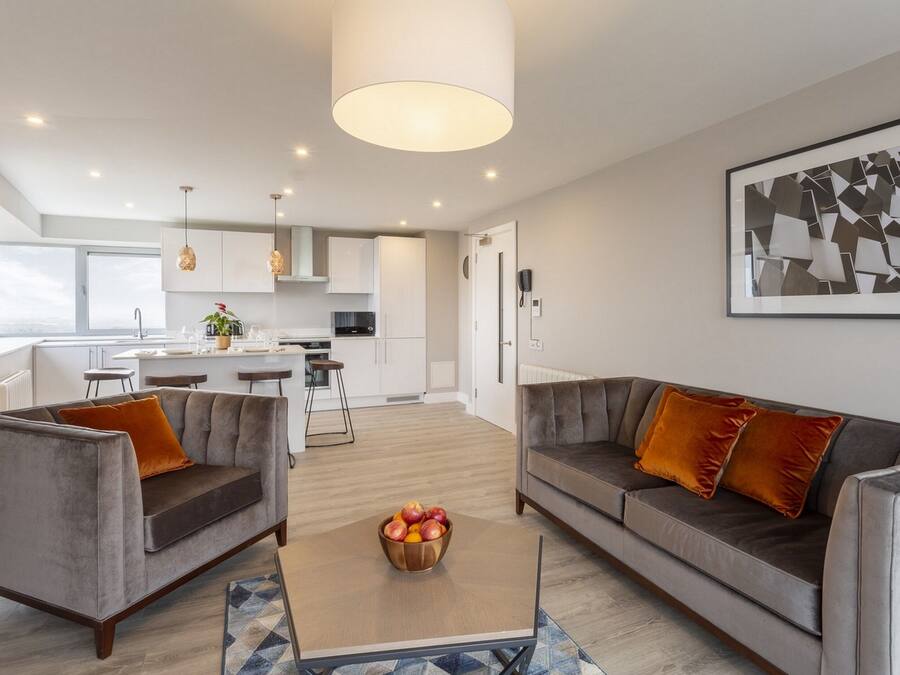 Een moderne woonkamer met een grijze bank, een salontafel met fruit, en een keuken op de achtergrond.