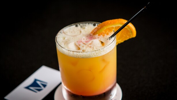 Un verre contenant une boisson de couleur orangée, agrémenté d’une tranche d’orange et d’une paille, posé sur un dessous de verre orné d’un logo.