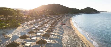 A beach with rows of sun loungers and thatched umbrellas, a lifeguard tower, and a clear view of the sea and distant hills.