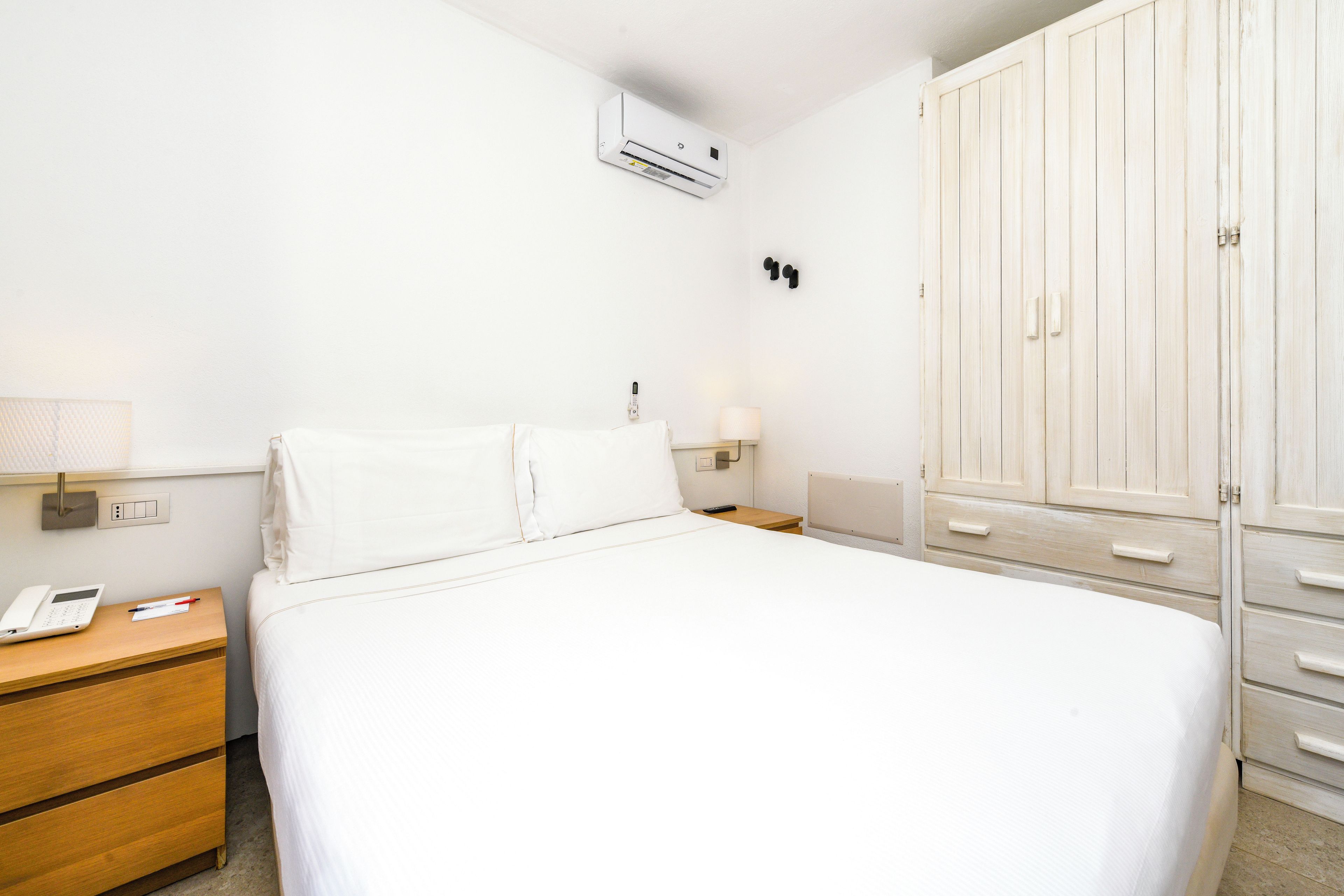 A hotel room with a bed, a nightstand, a lamp, a wardrobe, and an air conditioning unit.