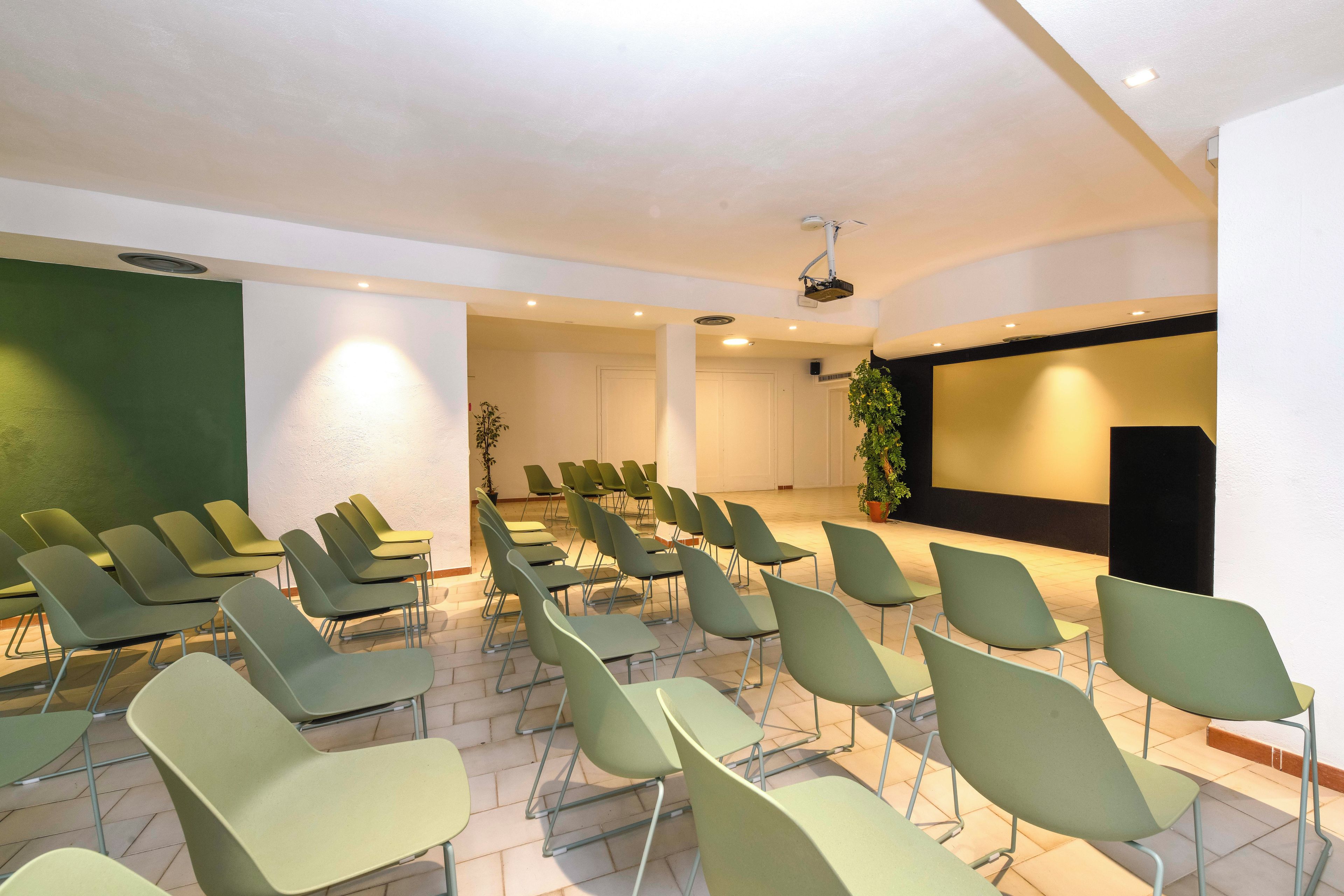 Un salón de conferencias con filas de sillas, un escenario con un orador y un proyector.