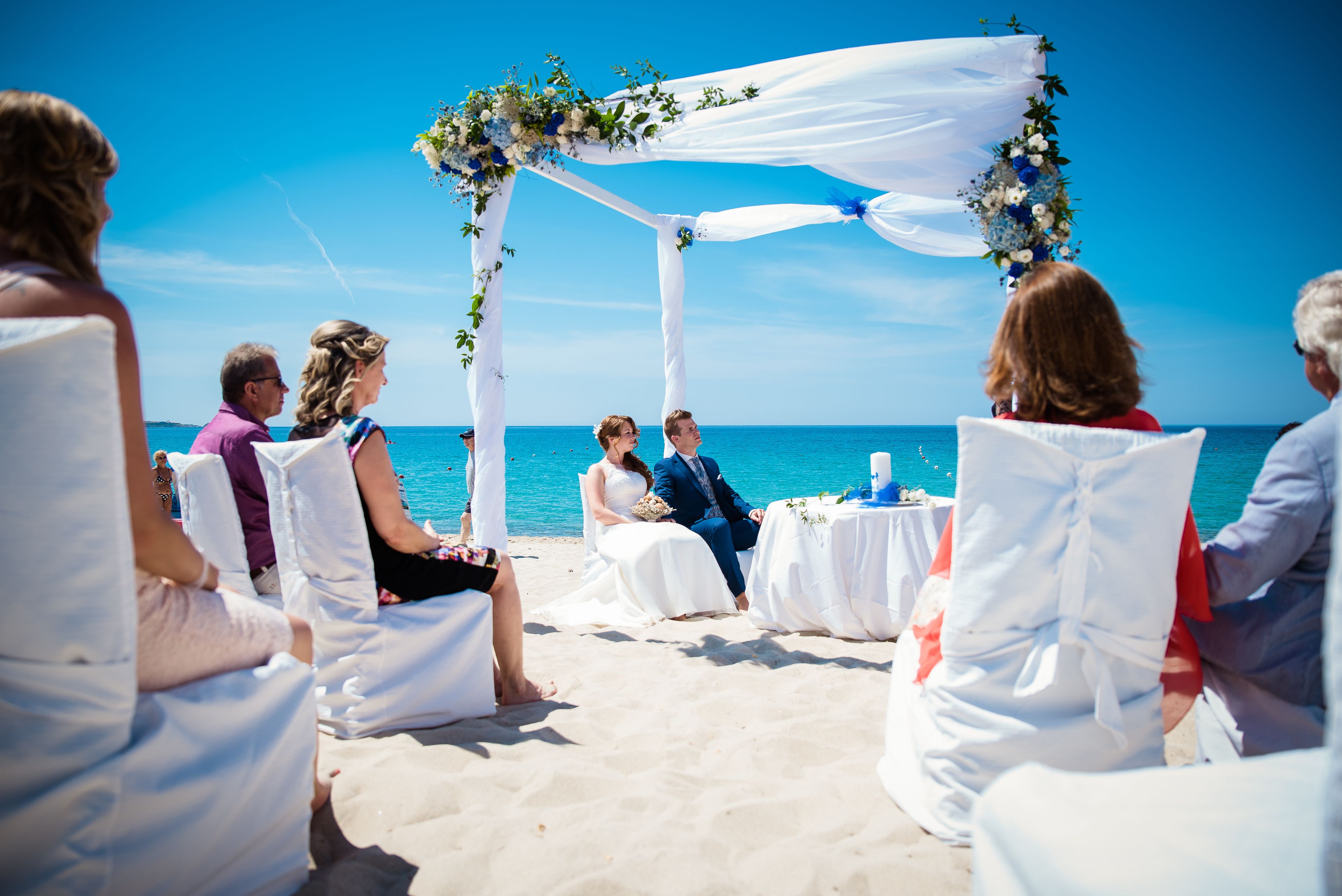 A beach wedding ceremony with white chairs and a floral arch.