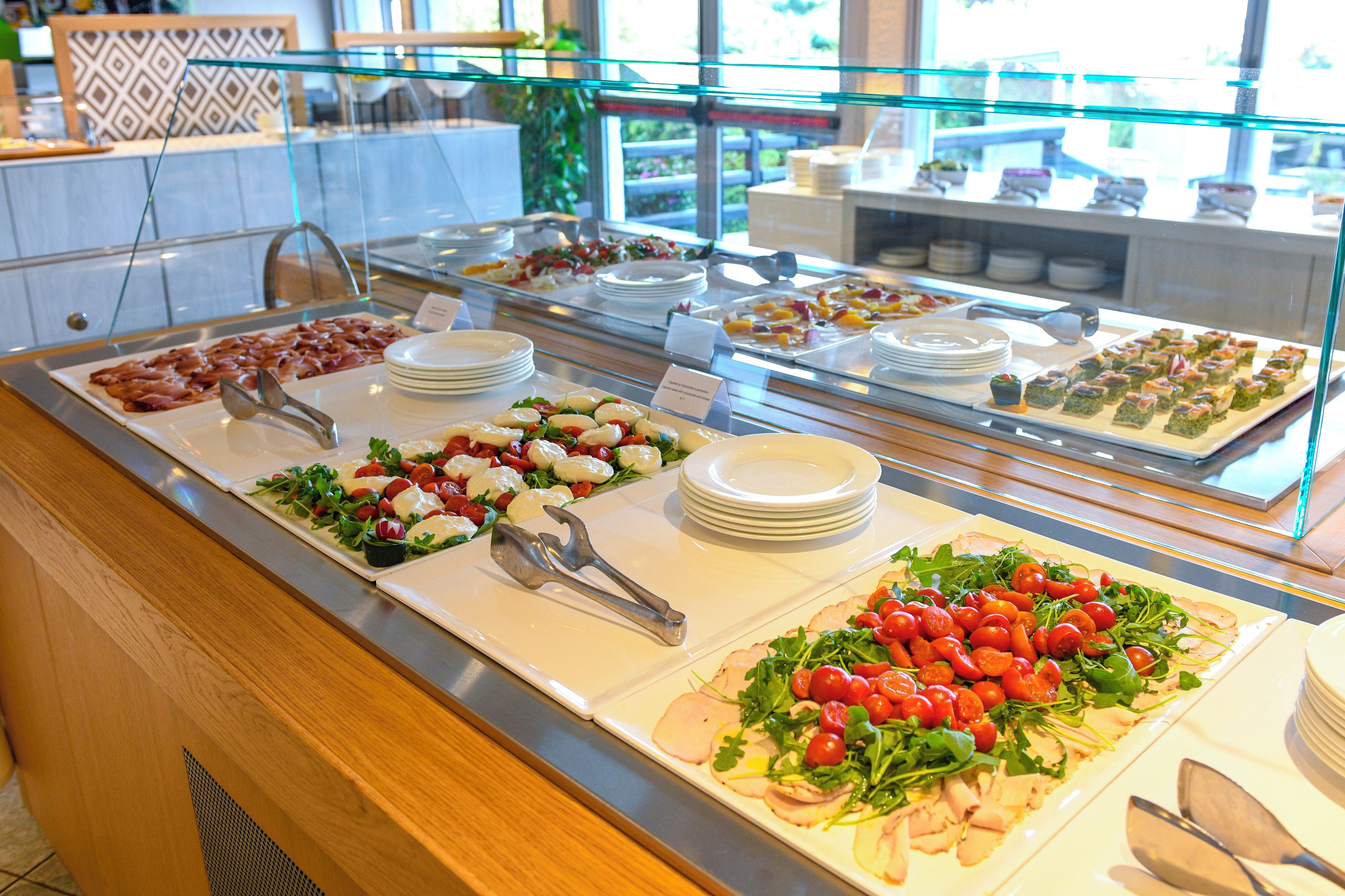 Un bufé con diversas opciones de comida, incluyendo ensaladas, carnes y postres, expuestos en un mostrador de vidrio.