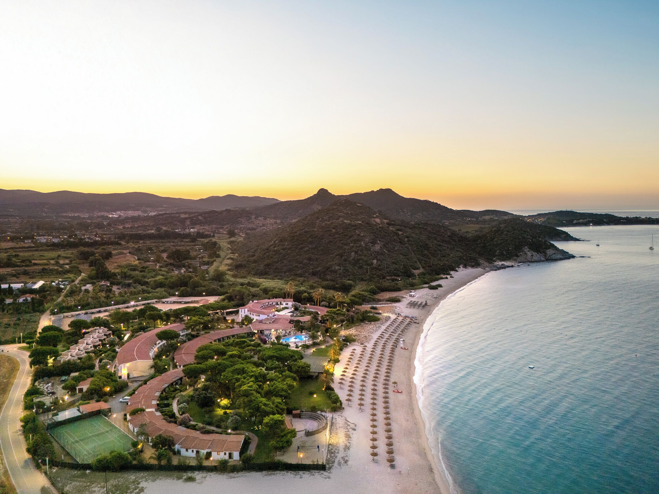 An aerial view of a coastal resort with a beach, tennis courts, and a mountainous backdrop.