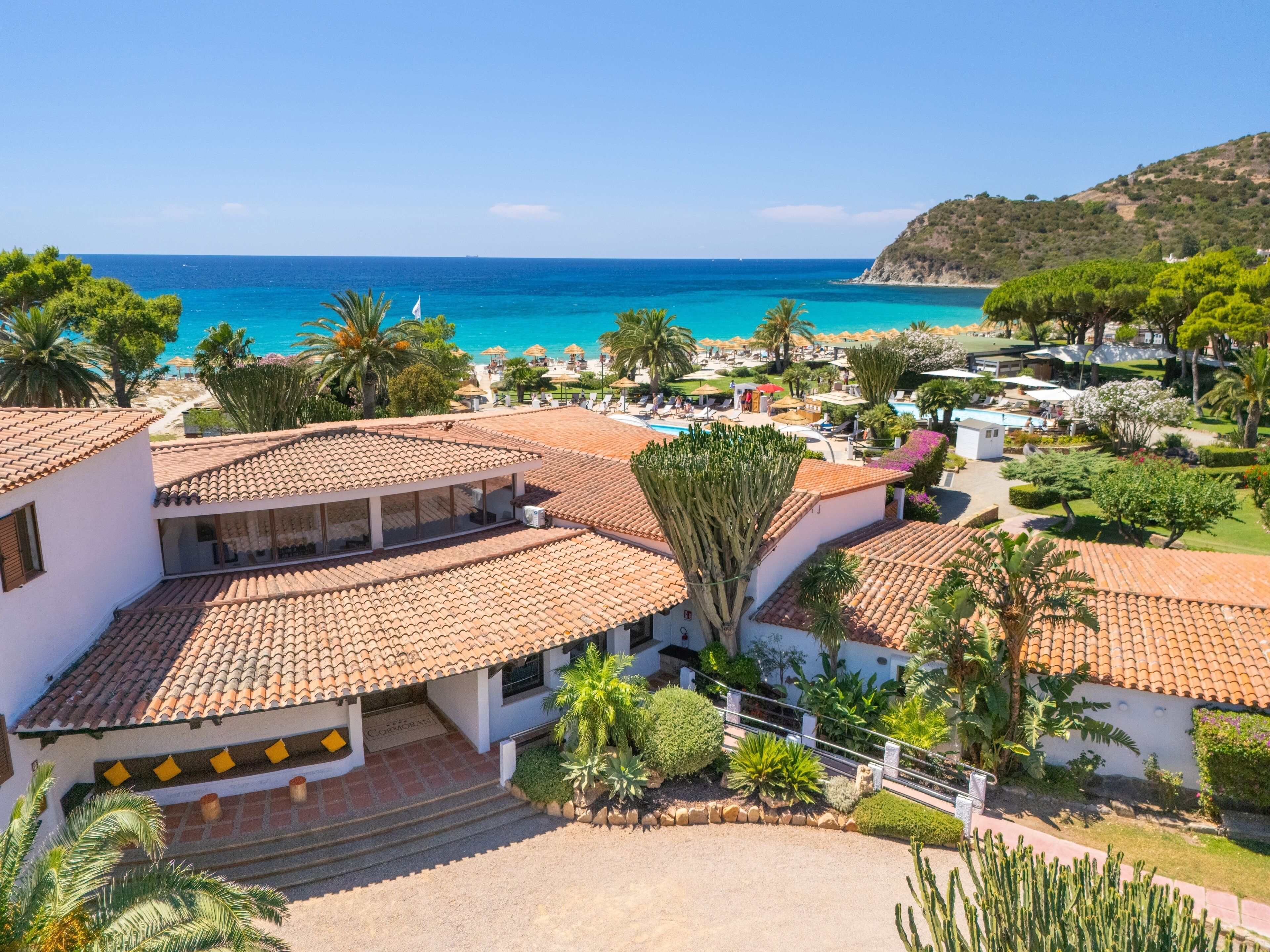A coastal resort with terracotta-tiled buildings, palm trees, and a clear view of the sea.