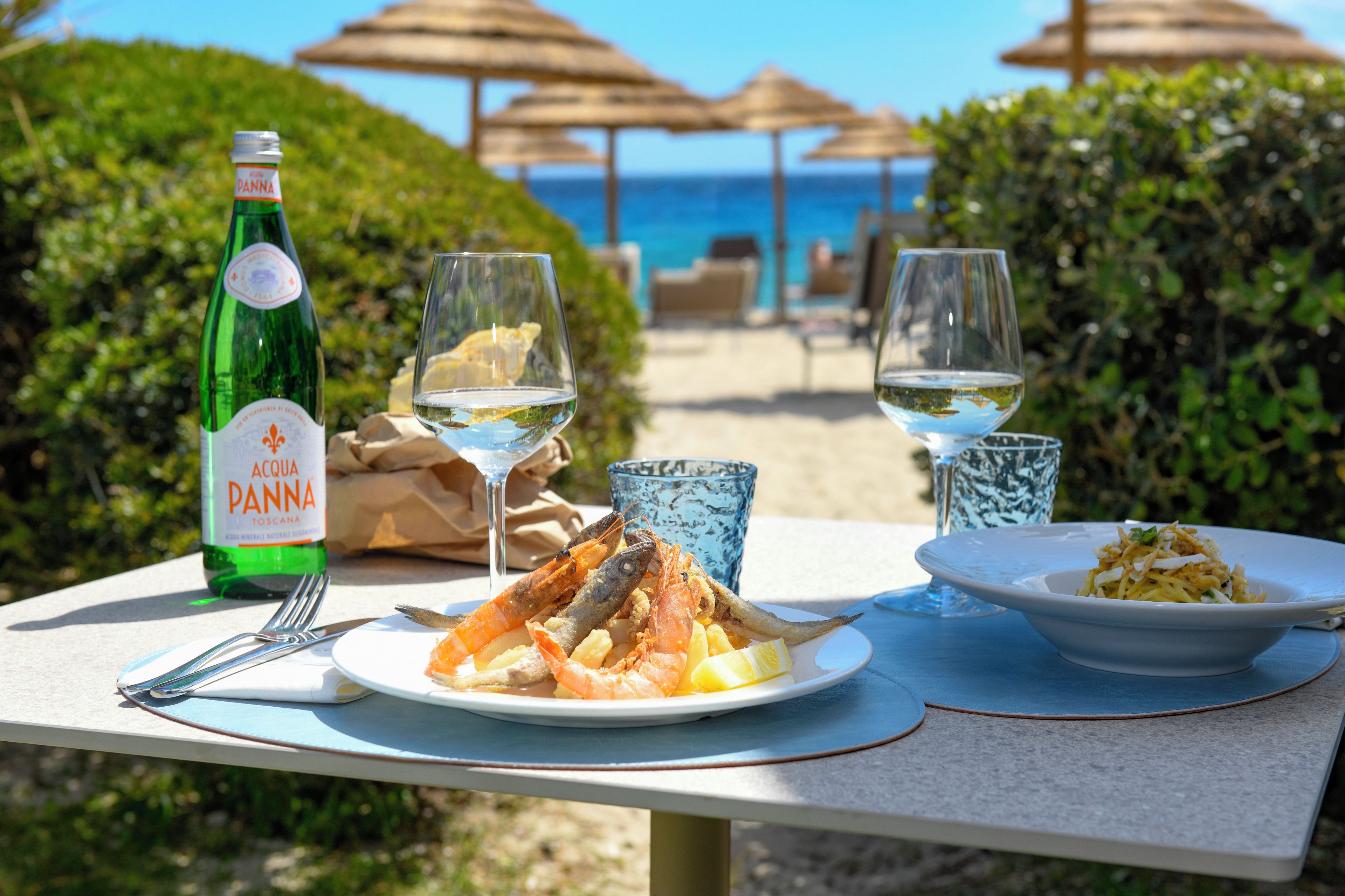 Uma mesa posta com comida, bebidas e uma garrafa de vinho Acqua Panna, com uma praia e guarda-sóis ao fundo.