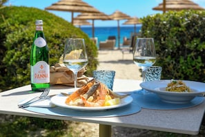 Una mesa dispuesta con comida, bebidas y una botella de vino Acqua Panna, con una playa y sombrillas al fondo.