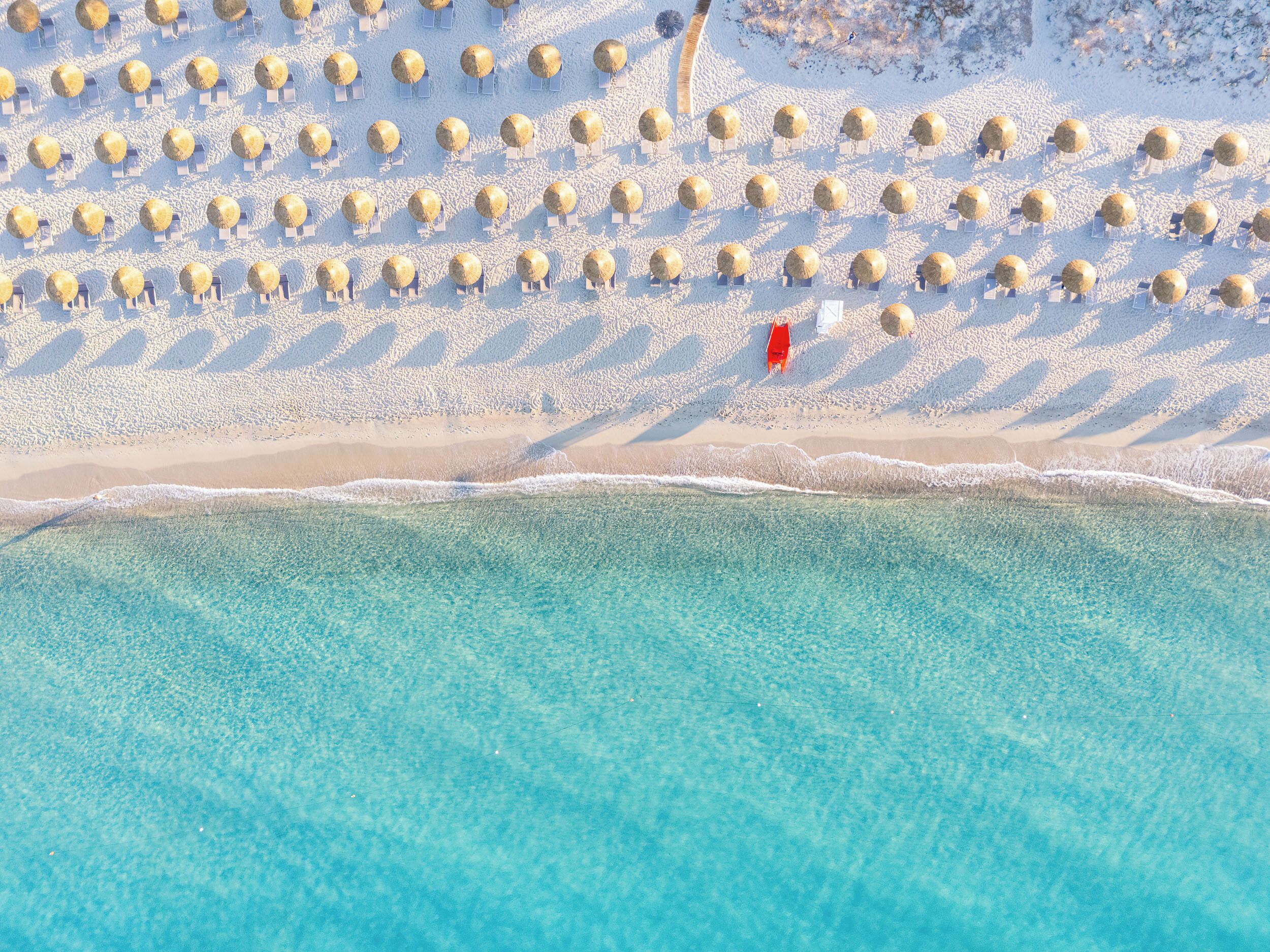 Vista aérea de una playa con hileras de sillas y tumbonas, orilla arenosa y agua turquesa cristalina.