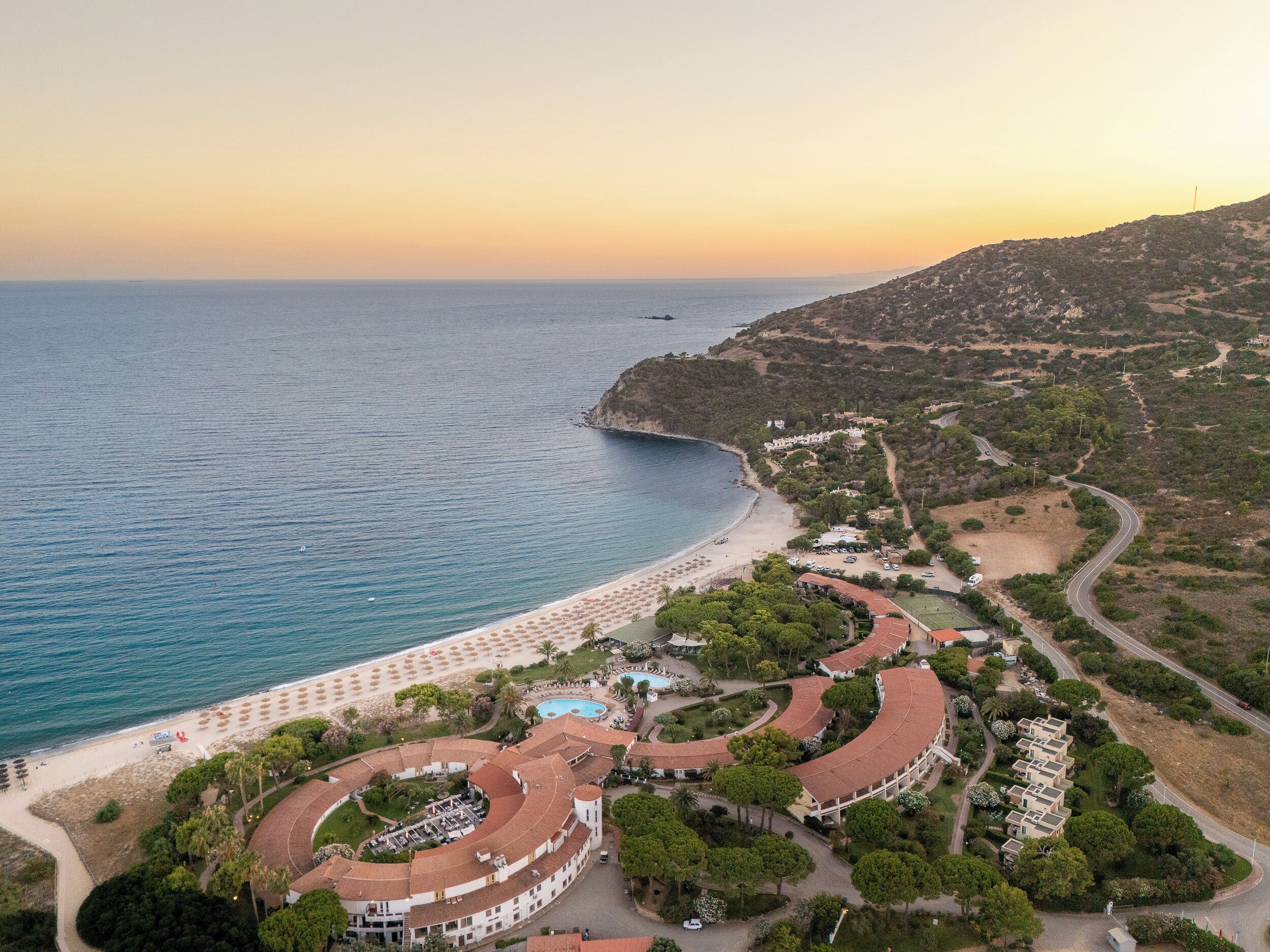 Ein Luftbild eines Küstenresorts mit einem gebogenen Gebäude, einem Strand mit Sonnenschirmen und freiem Blick auf das Meer.