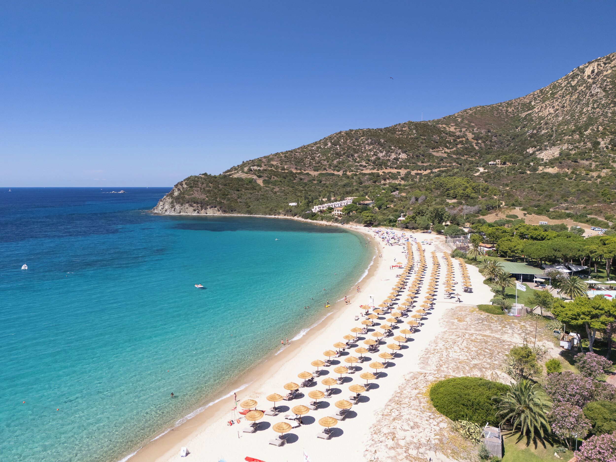 Une plage bordée de rangées de transats et de parasols, une mer d’un bleu limpide et un paysage montagneux en arrière-plan.