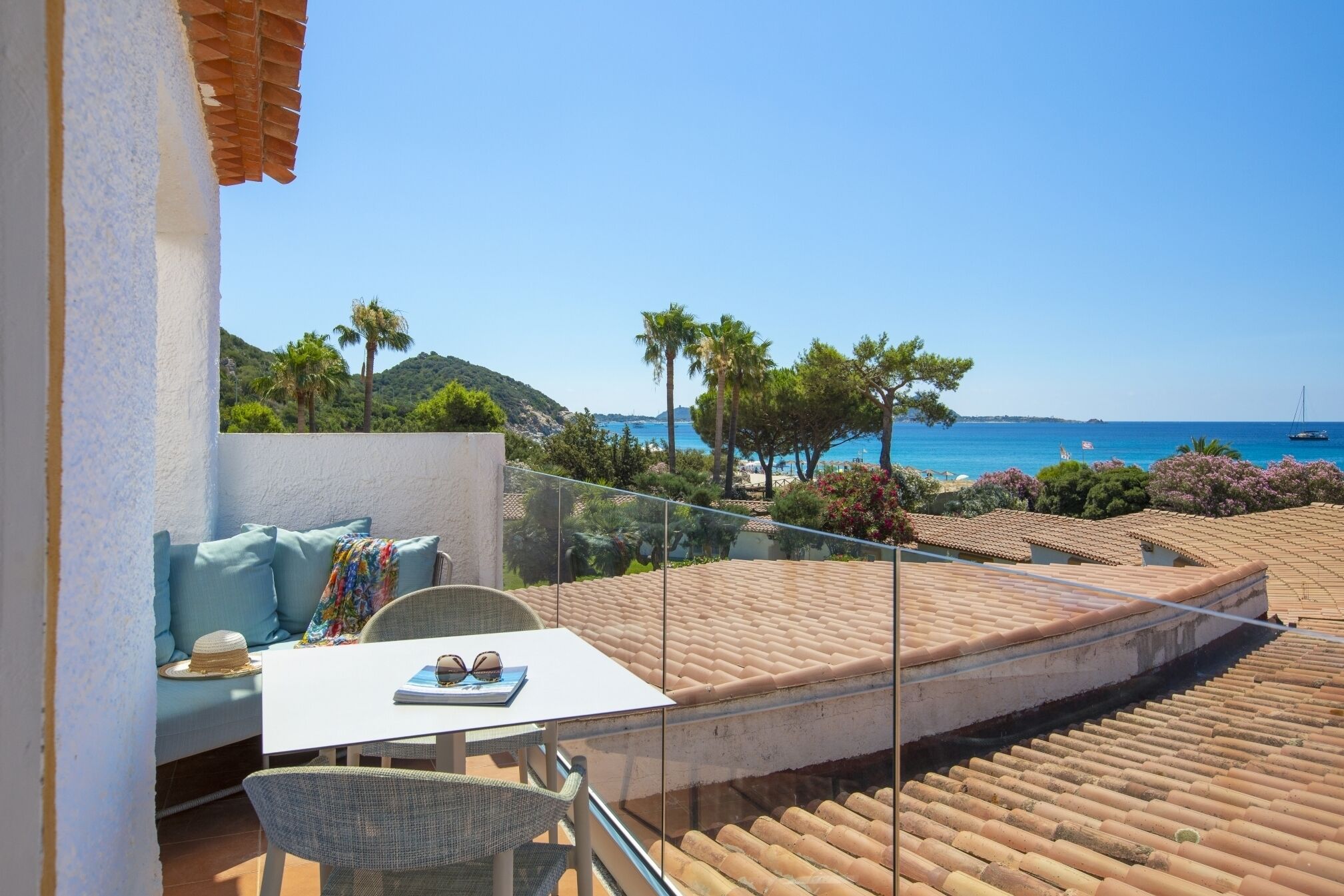 Un balcon doté d’une balustrade en verre donnant sur une plage, une table blanche avec un chapeau et des lunettes de soleil, et un canapé bleu.