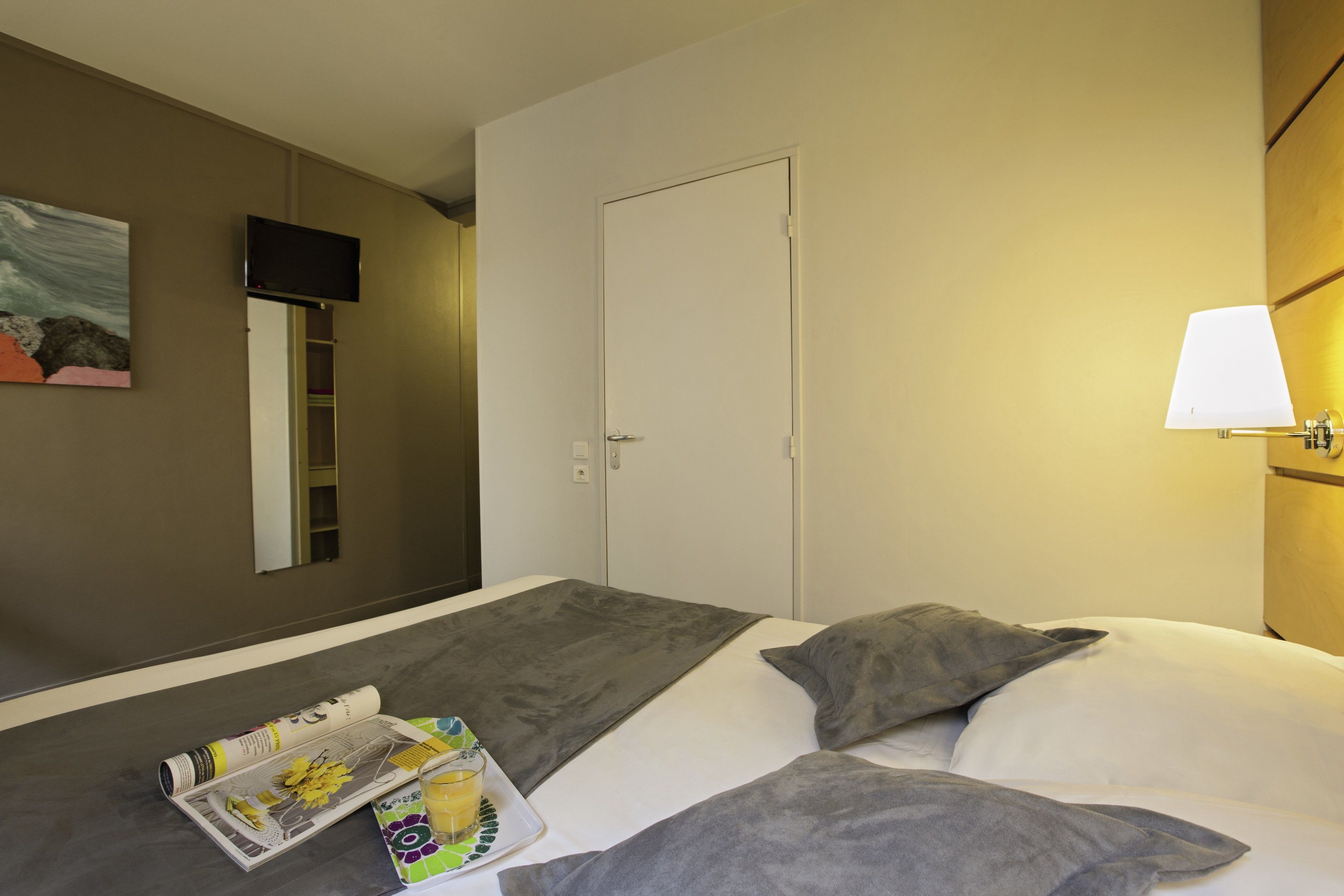 A hotel room with a bed, a flat-screen TV mounted on the wall, a door, and a bedside lamp.