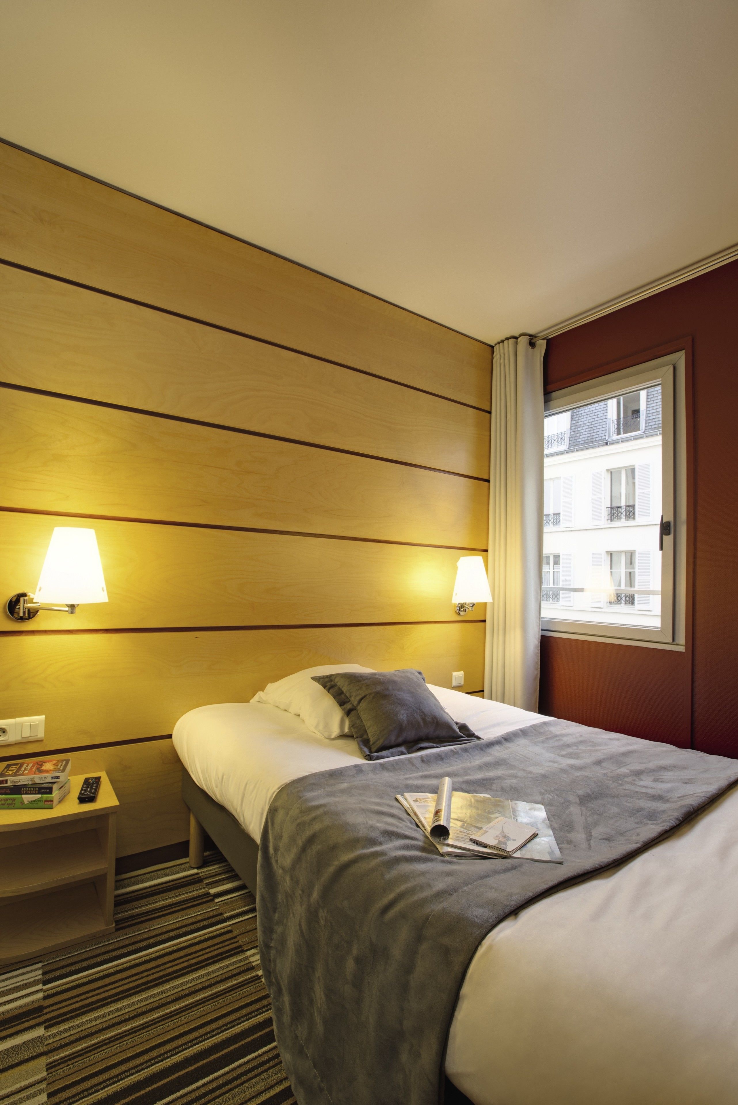 Una camera d'albergo con un letto, due lampade da comodino, un comodino con un telecomando e una finestra con vista sugli edifici.