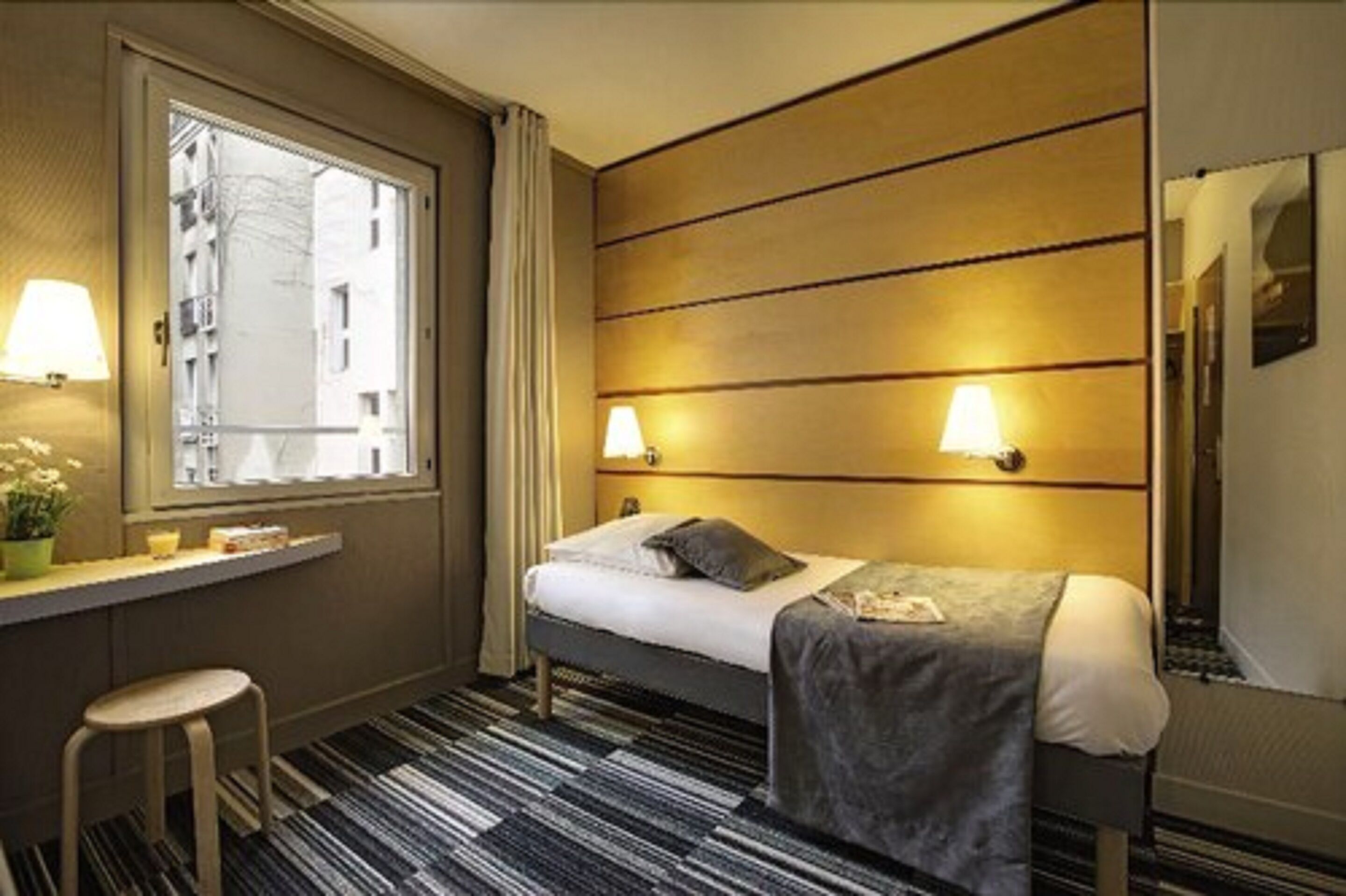 A hotel room with a single bed, bedside lamps, a window with curtains, a small table with a chair, and a striped carpet.