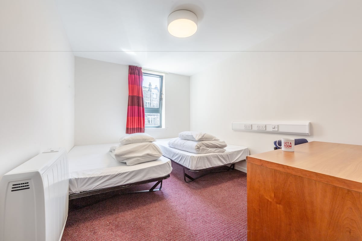 Une petite chambre propre, avec deux lits simples, un bureau en bois, un radiateur et une fenêtre ornée d’un rideau rouge.