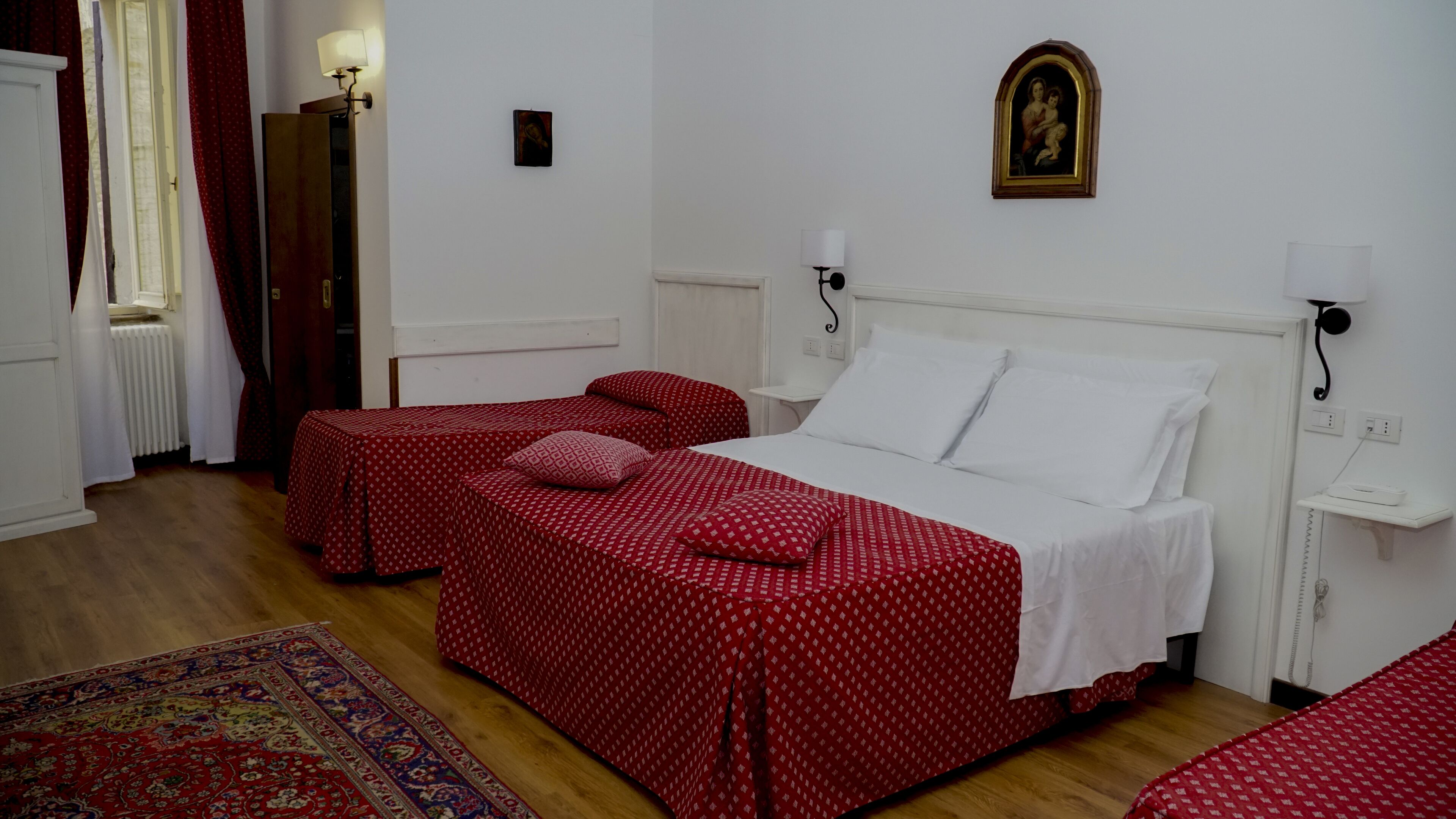 A room with a bed, two bedside tables, a nightstand, a radiator, a window with curtains, and a wall-mounted picture.