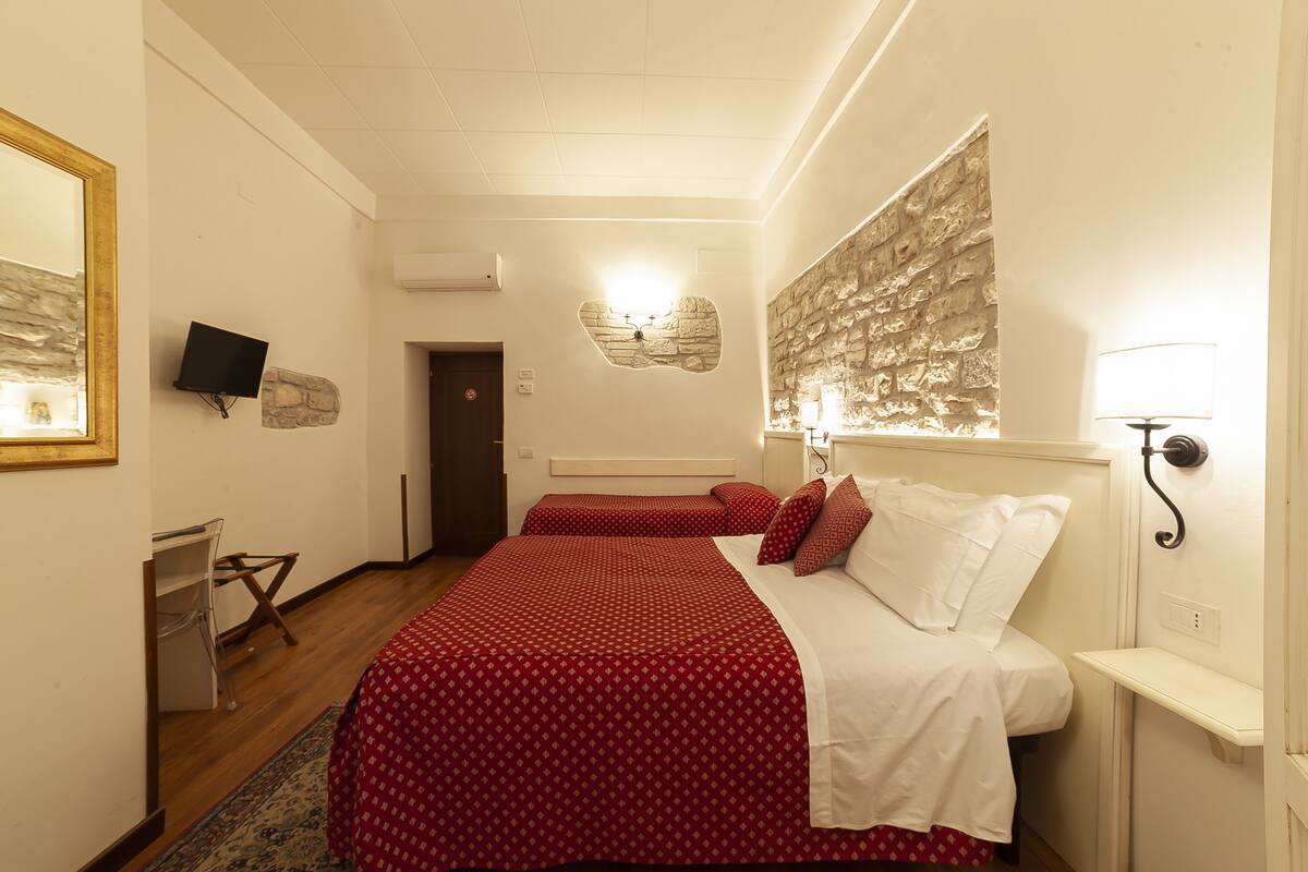 Quarto de hotel com duas camas, uma escrivaninha, uma televisão e uma parede de pedra.