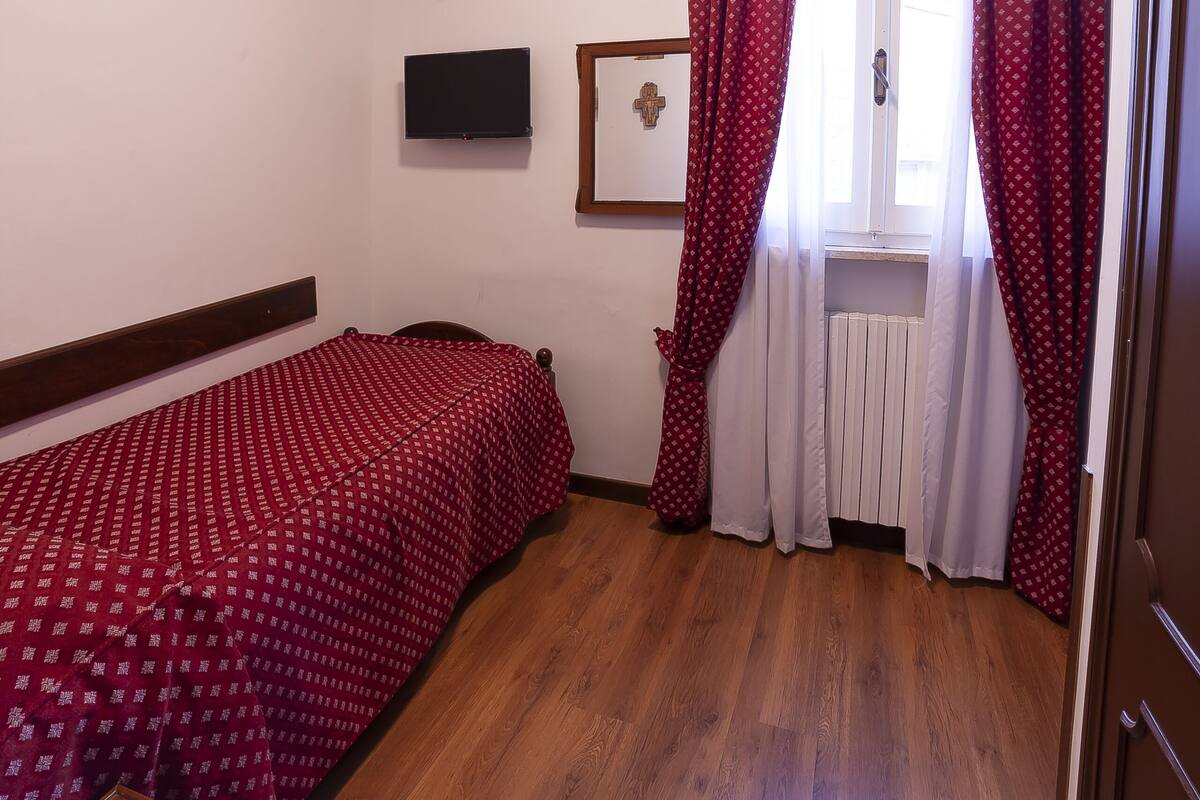 Quarto pequeno e arrumado com uma cama de solteiro, piso de madeira, janela com cortinas, televisão fixada na parede e radiador.