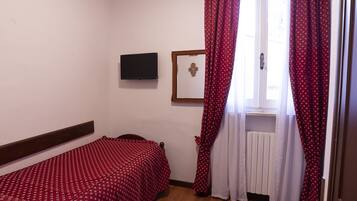 A small, tidy room with a single bed, a wooden floor, a window with curtains, a mounted TV, and a radiator.
