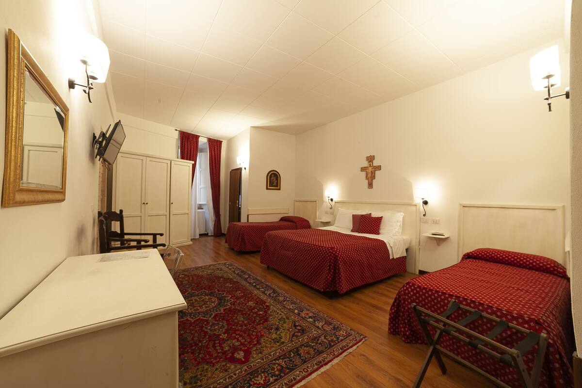 Quarto de hotel com duas camas de solteiro, uma escrivaninha, uma cadeira, um espelho, uma cruz na parede e um tapete vermelho estampado.