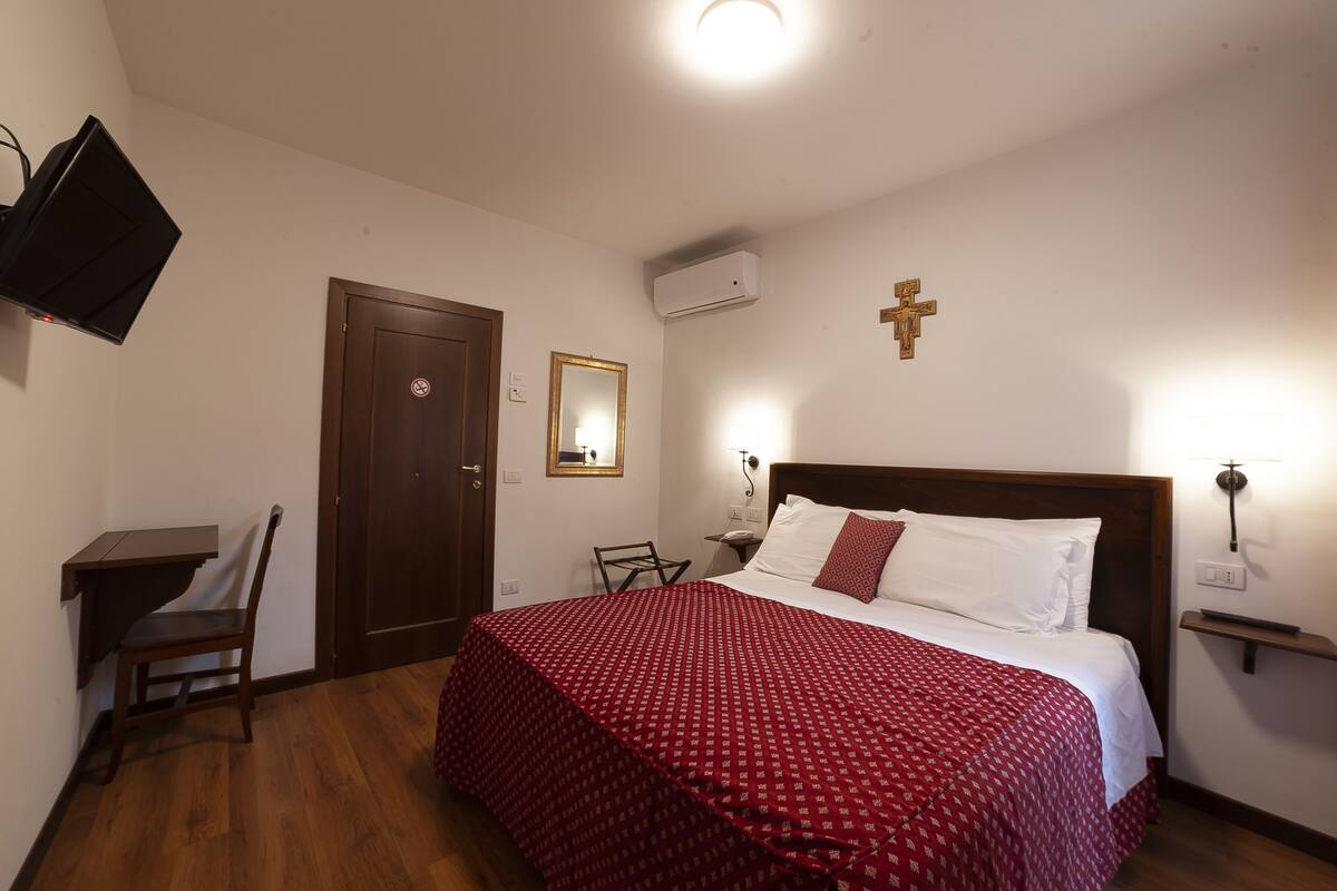 Quarto de hotel com uma cama, escrivaninha, cadeira, televisão, porta, ar condicionado e uma cruz pendurada na parede.