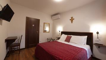A hotel room with a bed, a desk, a chair, a TV, a door, an air conditioning unit, and a wall-mounted cross.