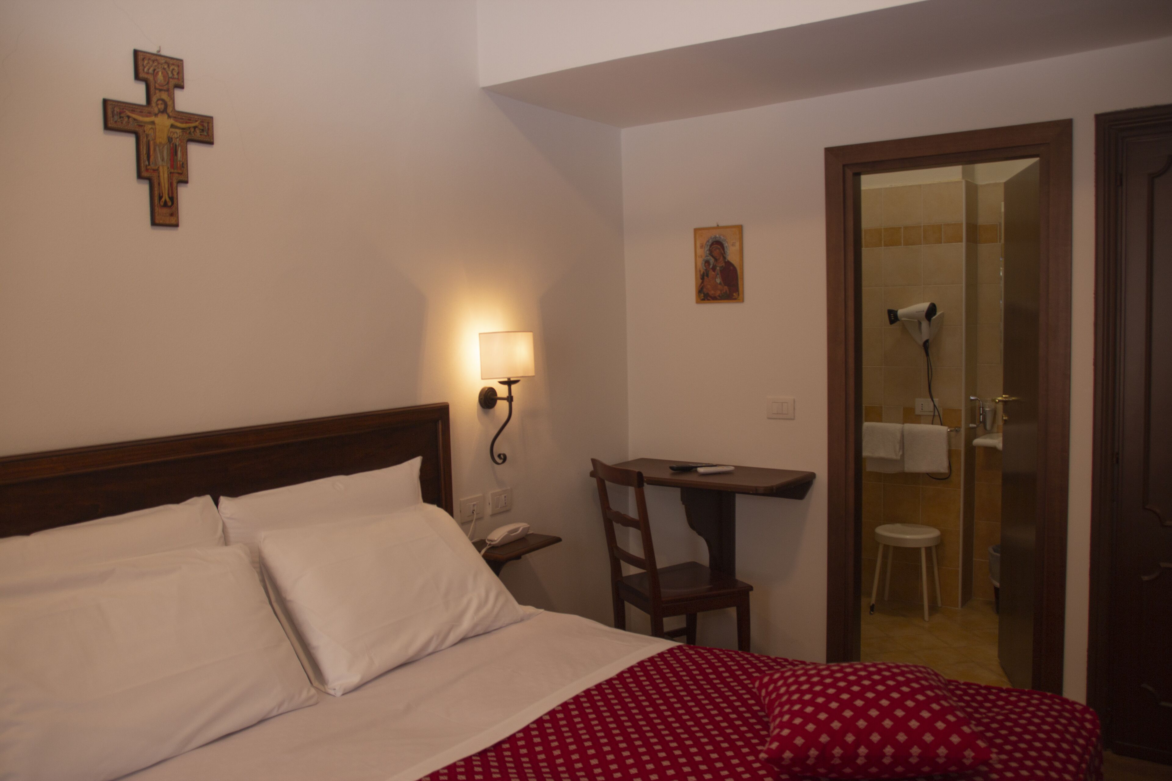 Quarto de hotel com cama, escrivaninha, cadeira, cruz na parede, luminária e um quadro.
