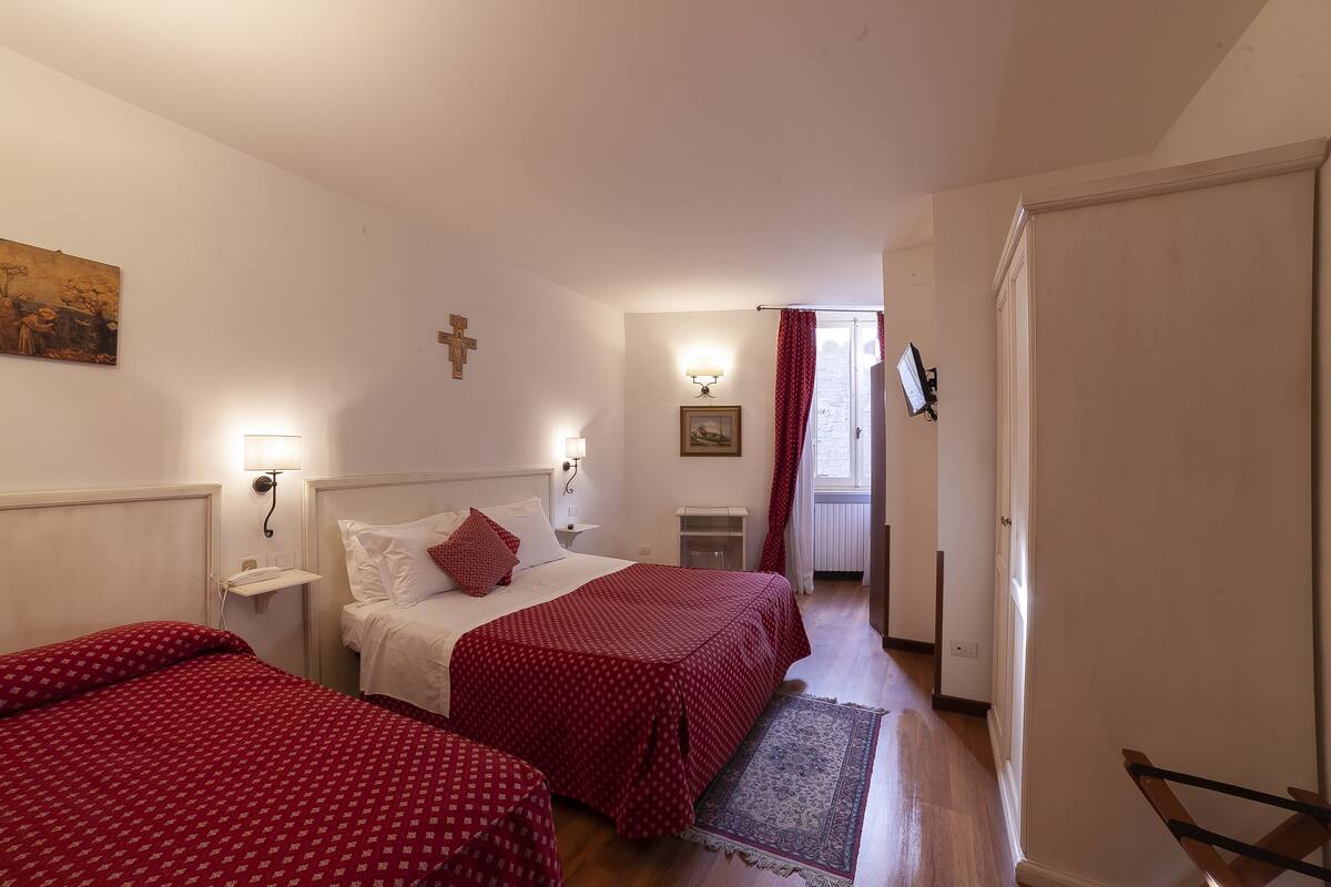 Quarto de hotel com duas camas, uma mesa de cabeceira, uma luminária na parede, um quadro emoldurado e uma janela com cortinas.