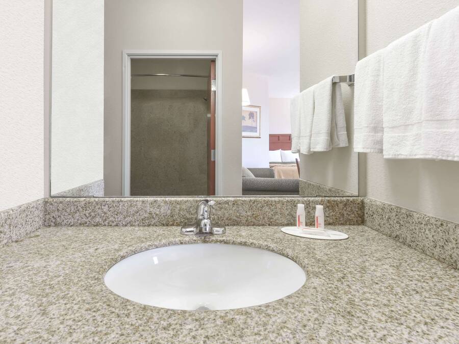 A bathroom with a granite countertop, a white sink, and a mirror reflecting a door and a bed.