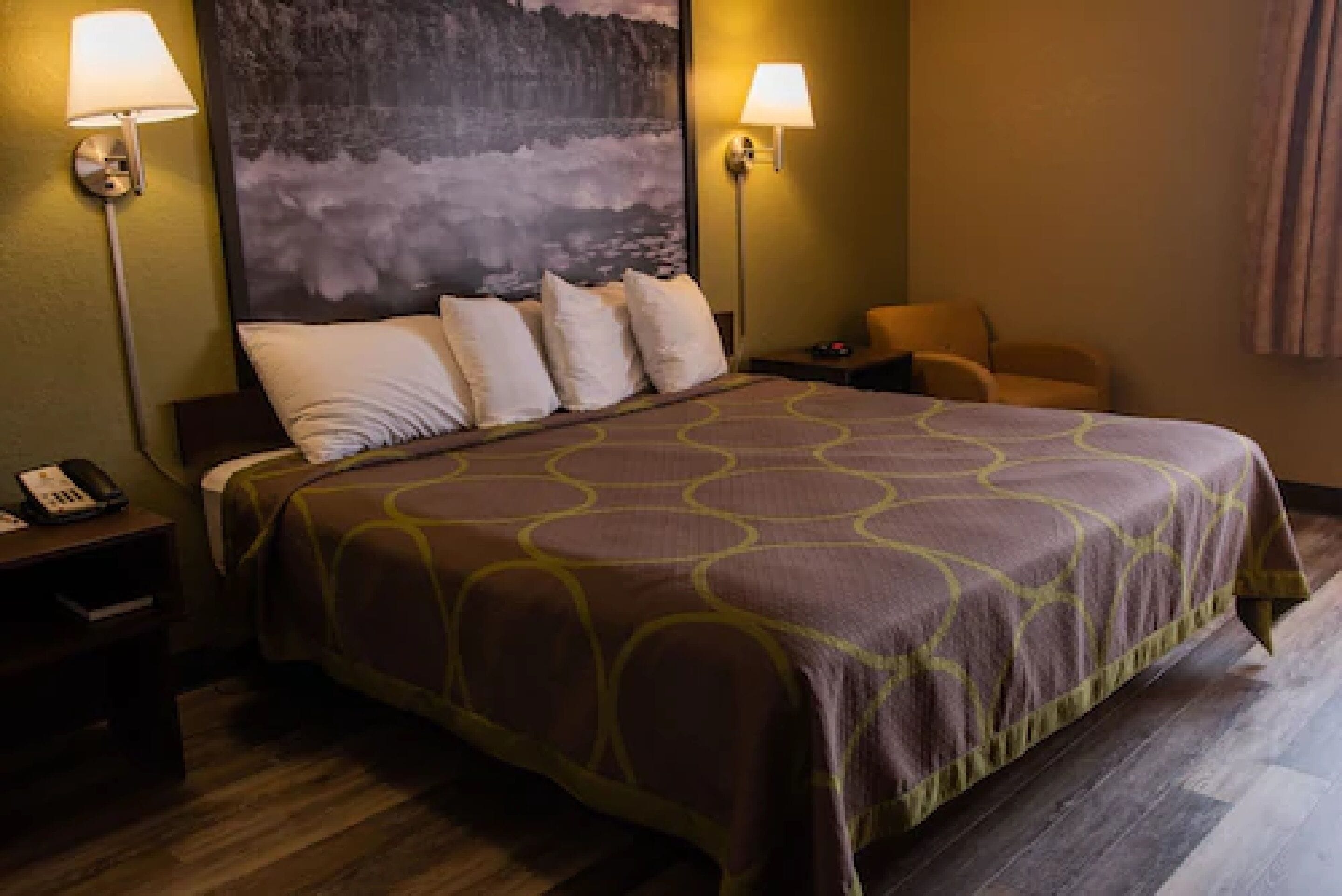 A hotel room with a large bed, two bedside lamps, a chair, and a painting on the wall.