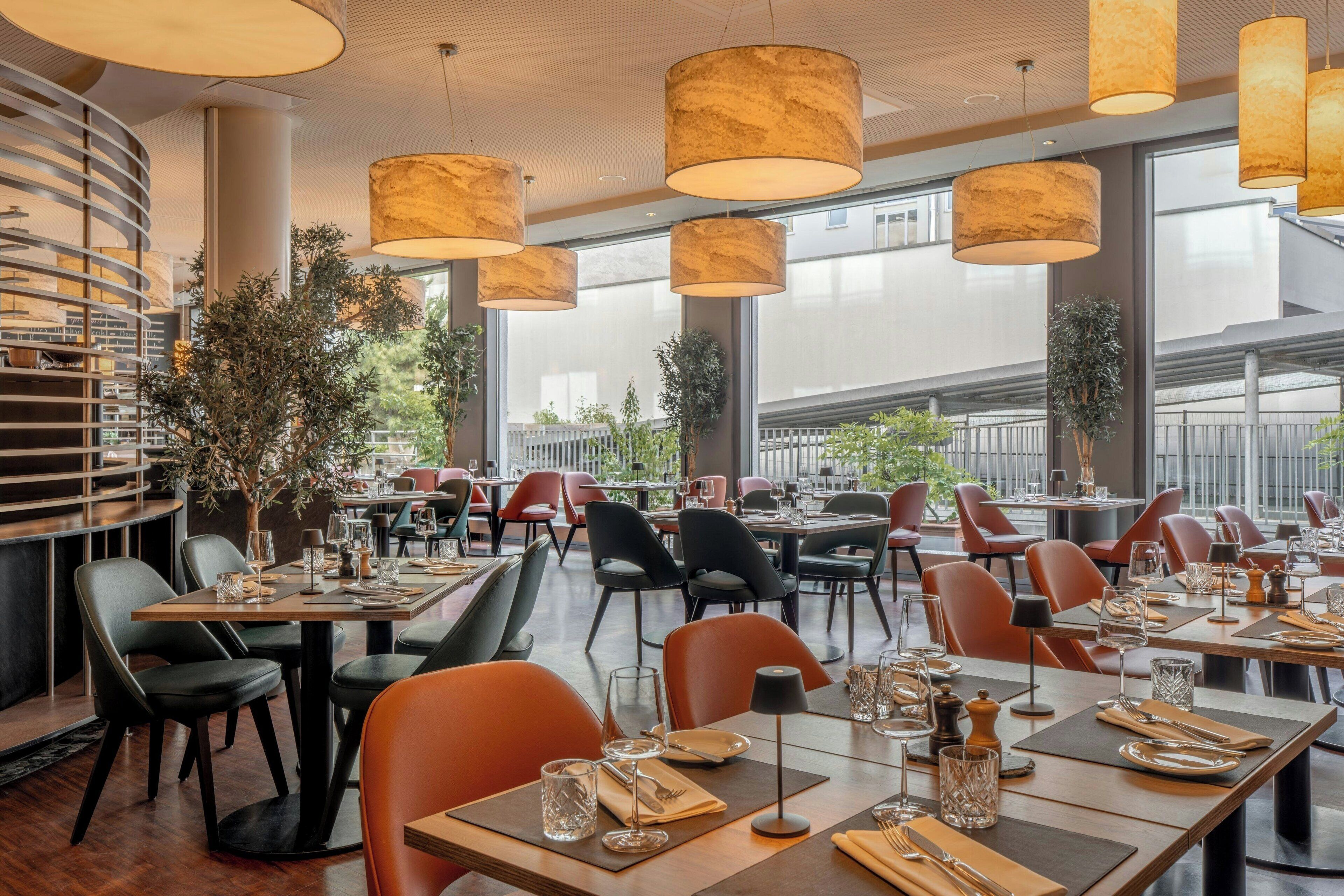Un restaurante con decoración moderna, con mesas redondas, sillas negras y anaranjadas, y lámparas colgantes grandes.