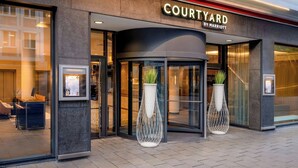 Entrada de un hotel Courtyard by Marriott con un diseño moderno, destacando puertas de cristal amplias, una entrada curva y dos macetas decorativas.