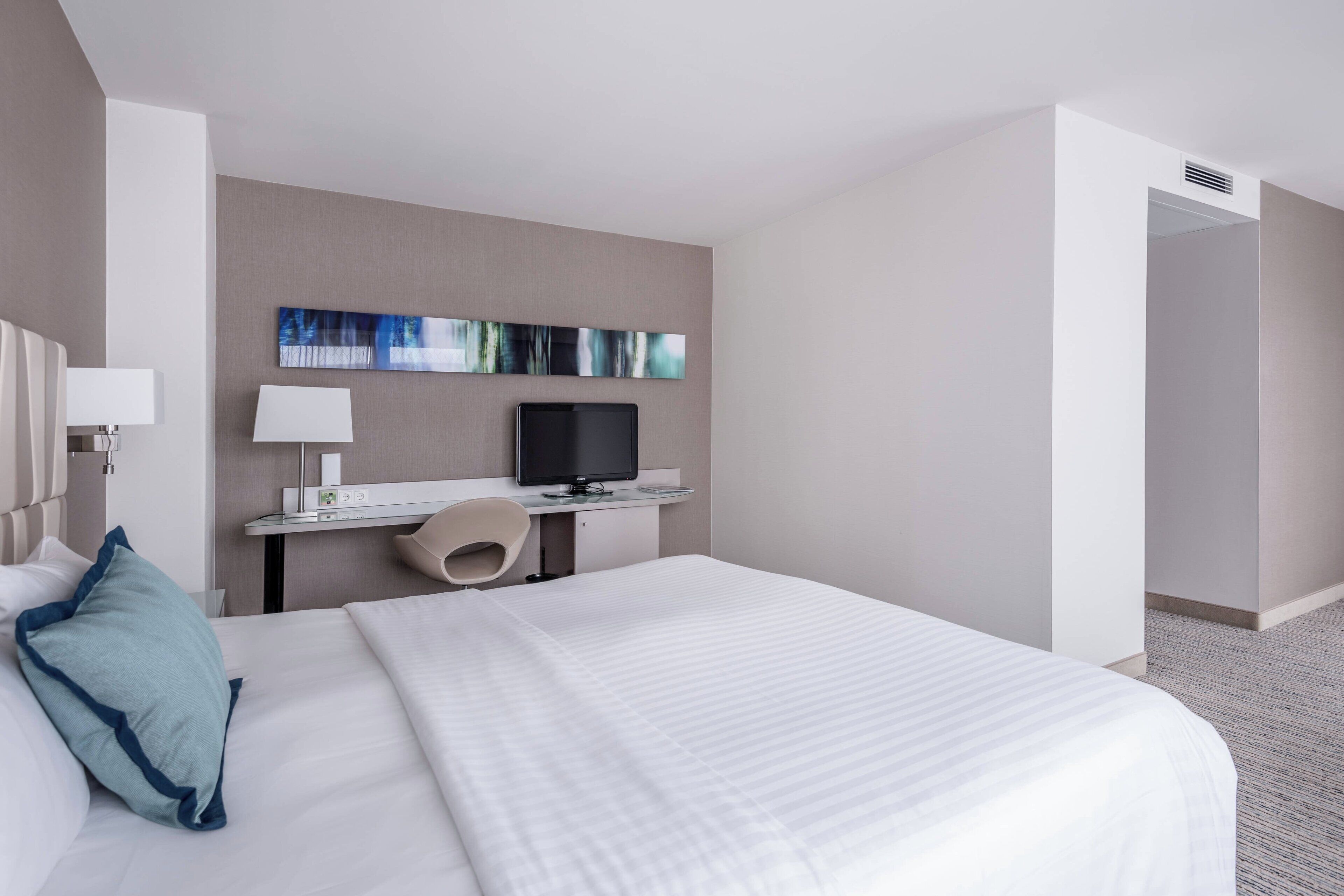 A hotel room with a large bed, a desk with a computer, a TV, and a wall-mounted artwork.