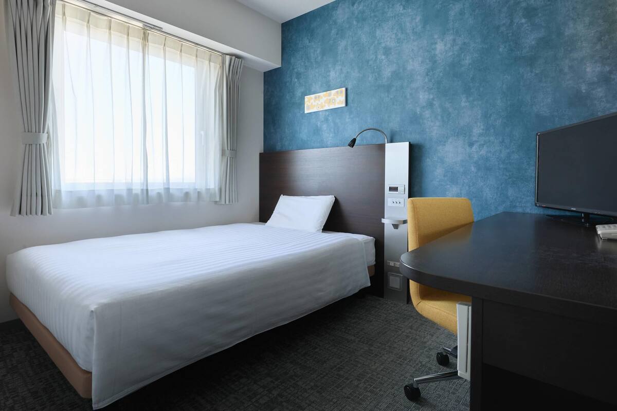 Une chambre d’hôtel équipée d’un lit, d’un bureau, d’une chaise, d’une télévision et d’un panneau de commande fixé au mur.
