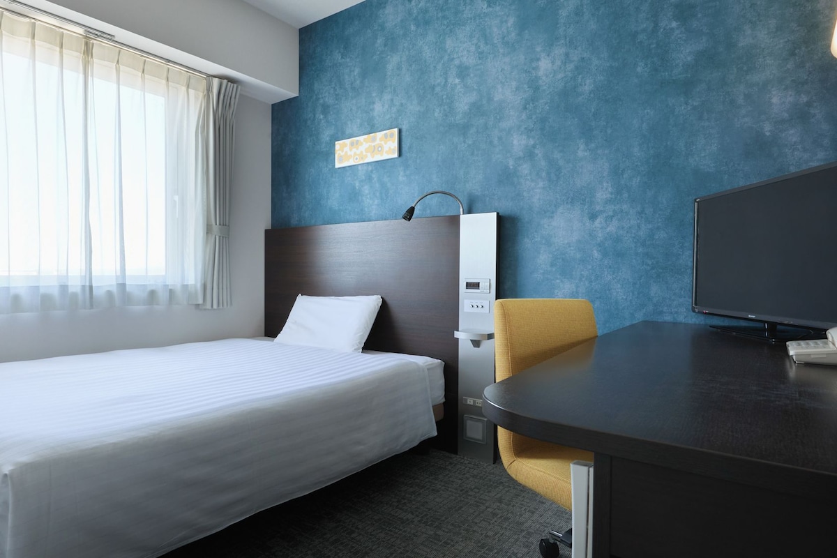 A hotel room with a bed, a desk with a television, a chair, and a wall-mounted heater.