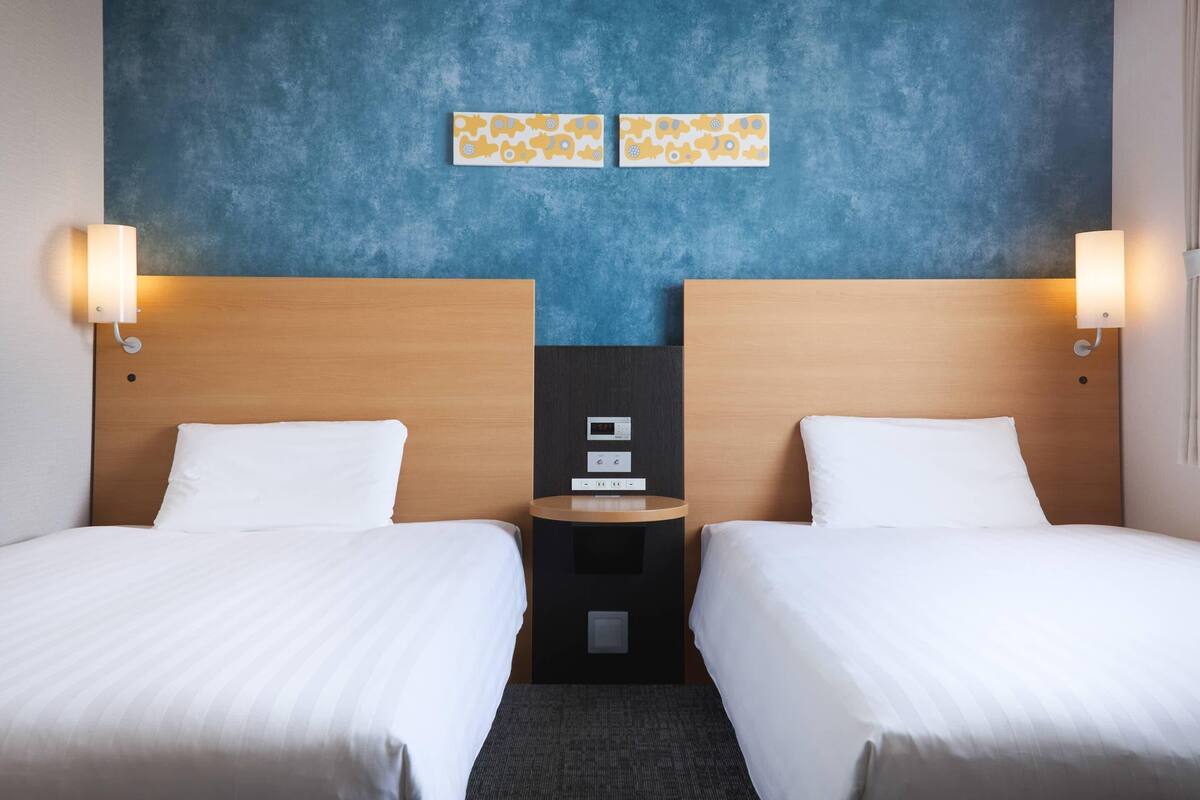 Une chambre d’hôtel avec deux lits, une table de chevet, une horloge murale et deux lampes fixées au mur.