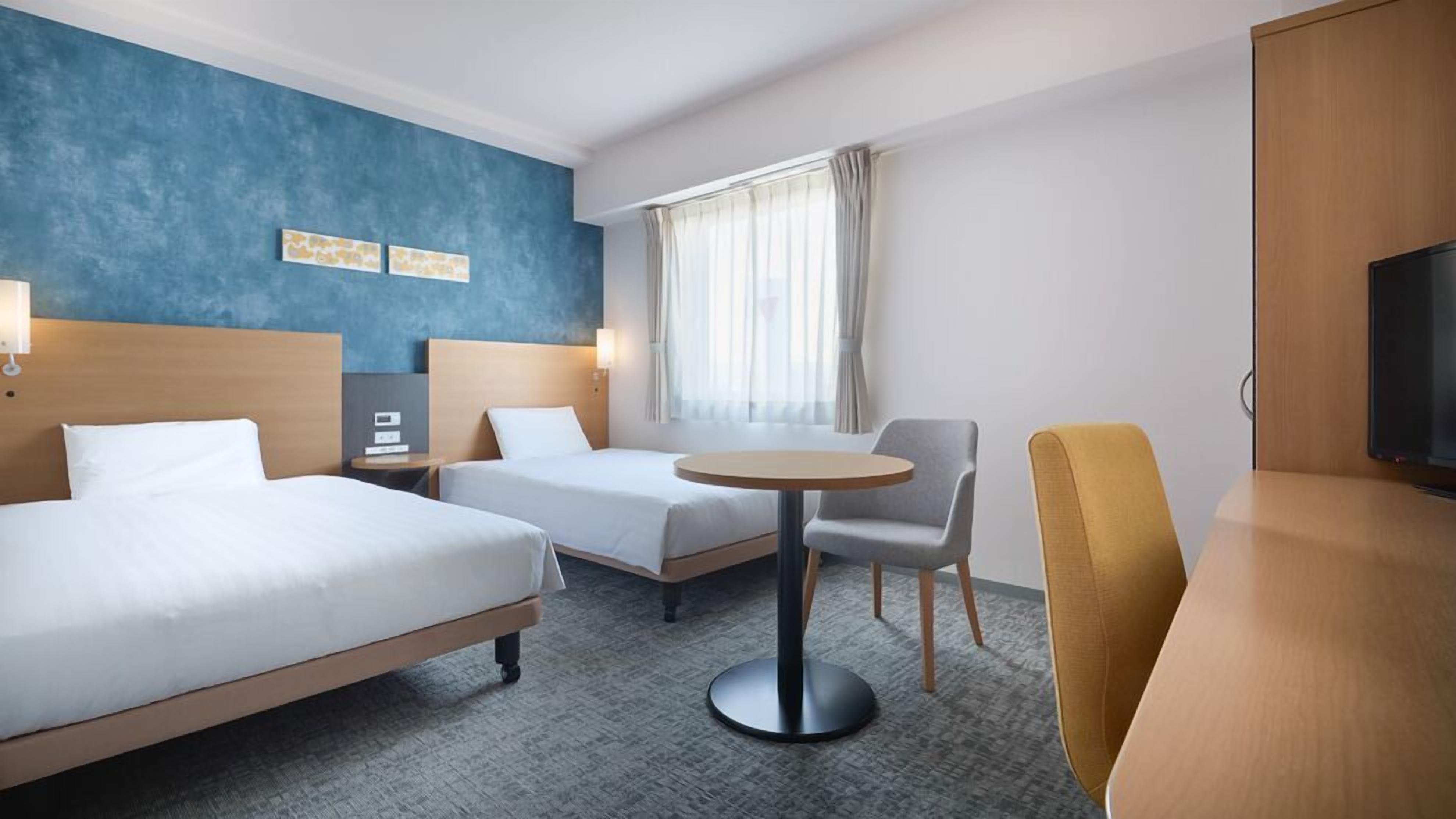 Una habitación de hotel con dos camas, una mesita redonda pequeña, un televisor y una ventana con cortinas.