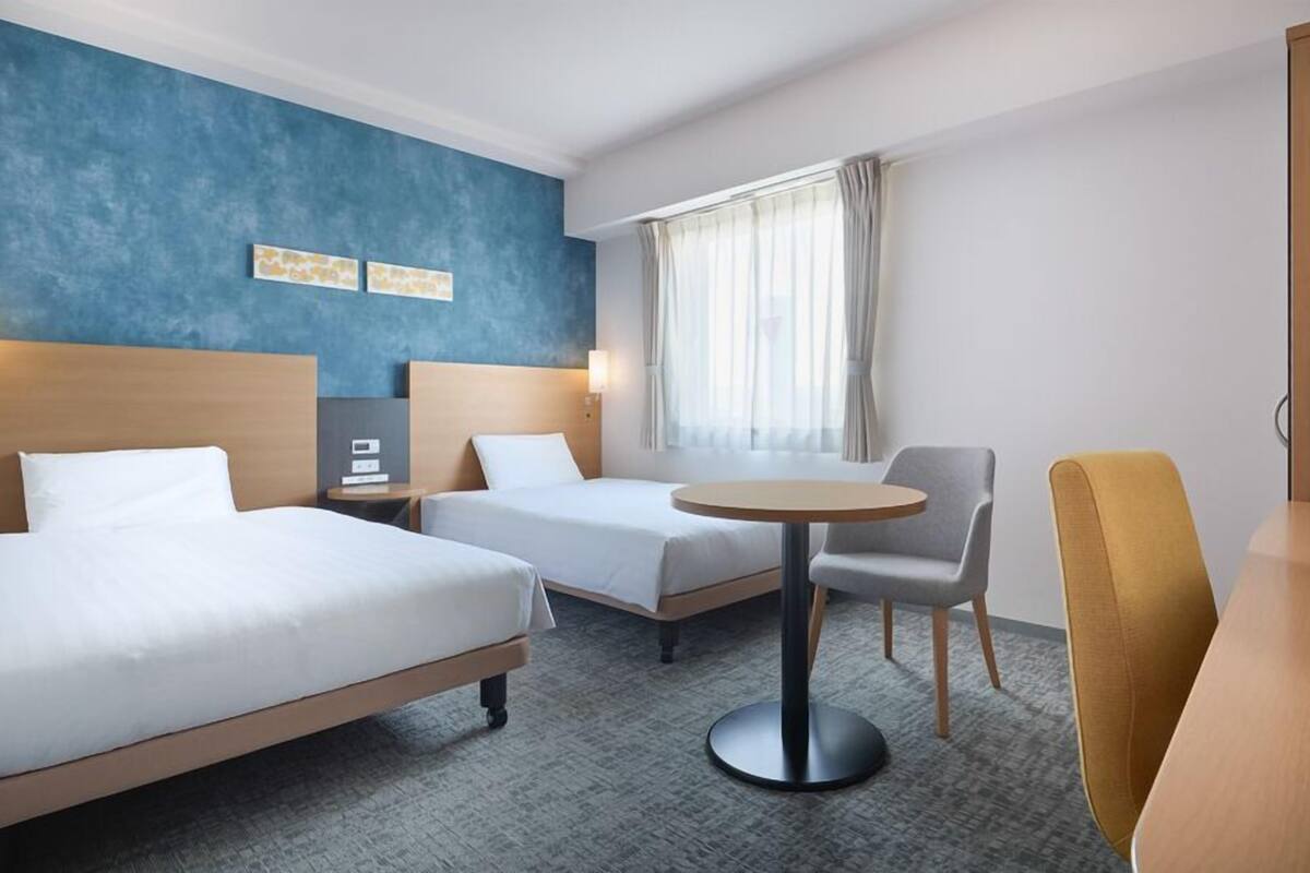 Una habitación de hotel con dos camas, una mesita redonda pequeña, un televisor y una ventana con cortinas.