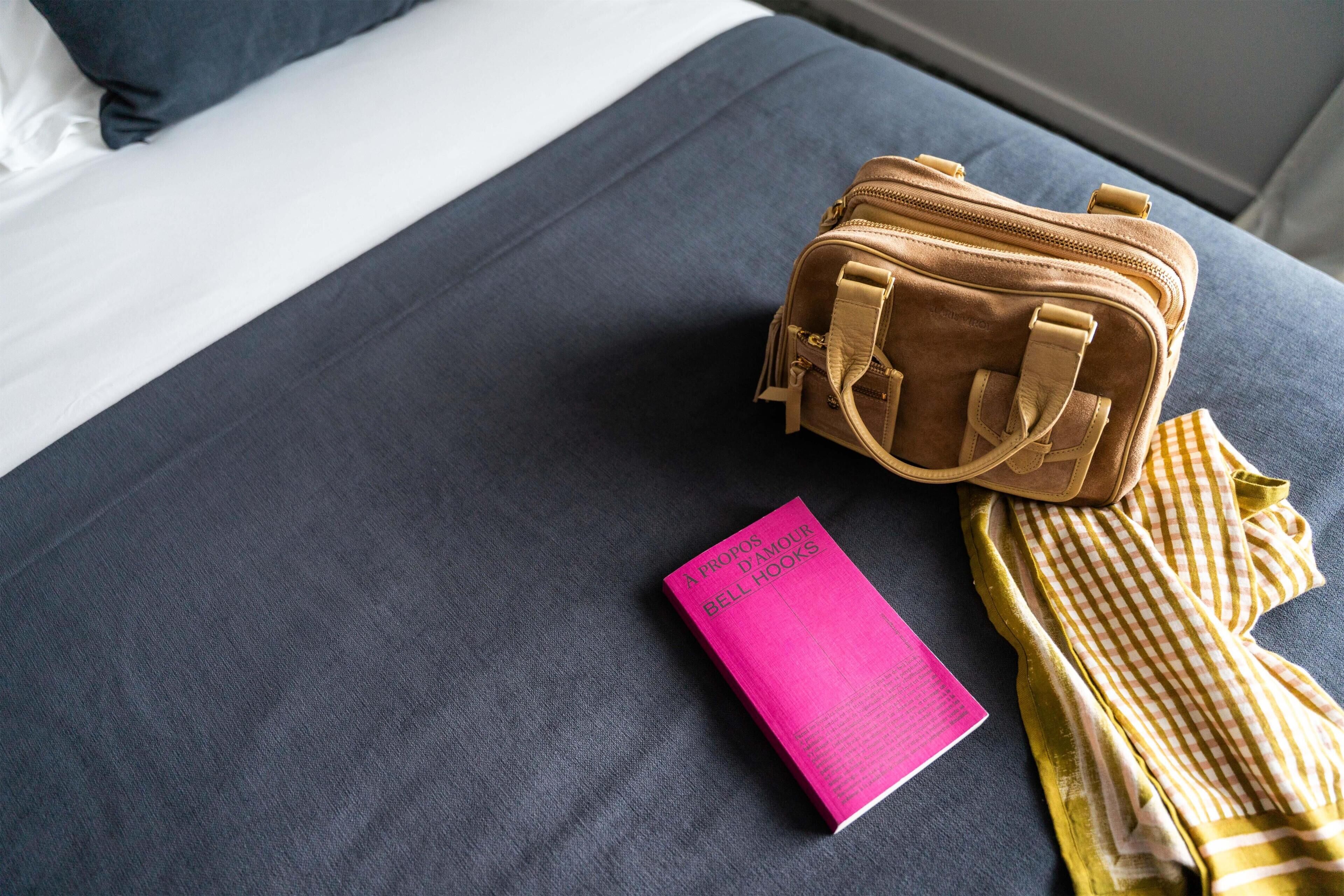 Una cama con una colcha azul oscuro, un bolso de cuero marrón, un libro rosa titulado 'A propos d'amour' y un paño amarillo a cuadros.