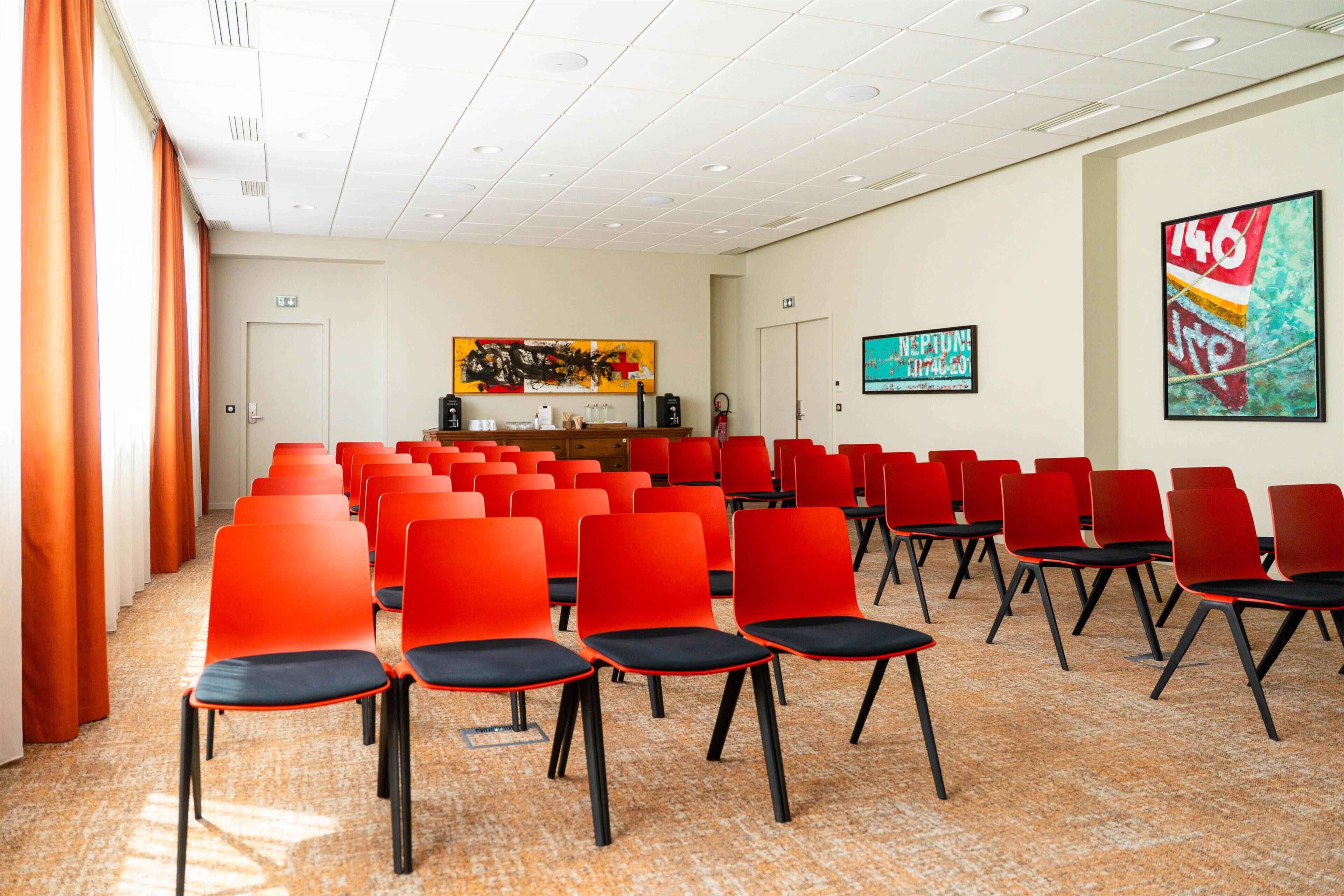 A conference room with rows of red chairs, a white ceiling, and artwork on the walls.