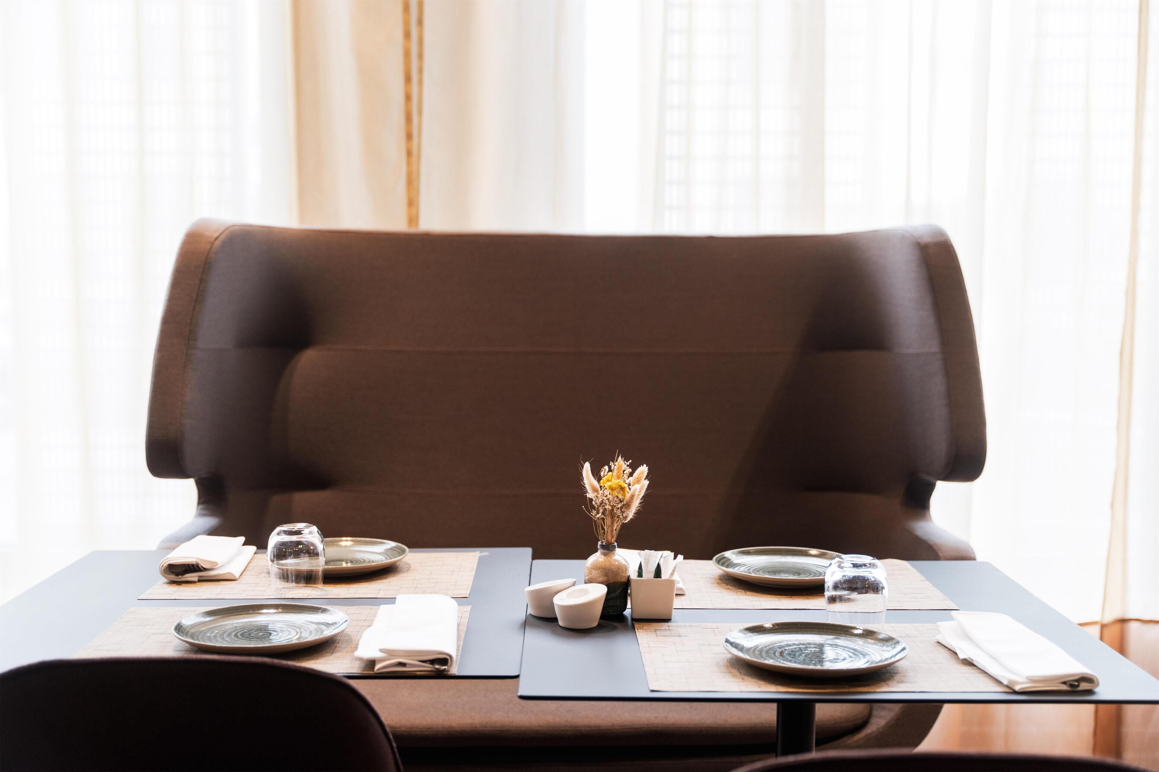 A dining table set with plates, glasses, and a vase with dried flowers, positioned in front of a brown upholstered chair.