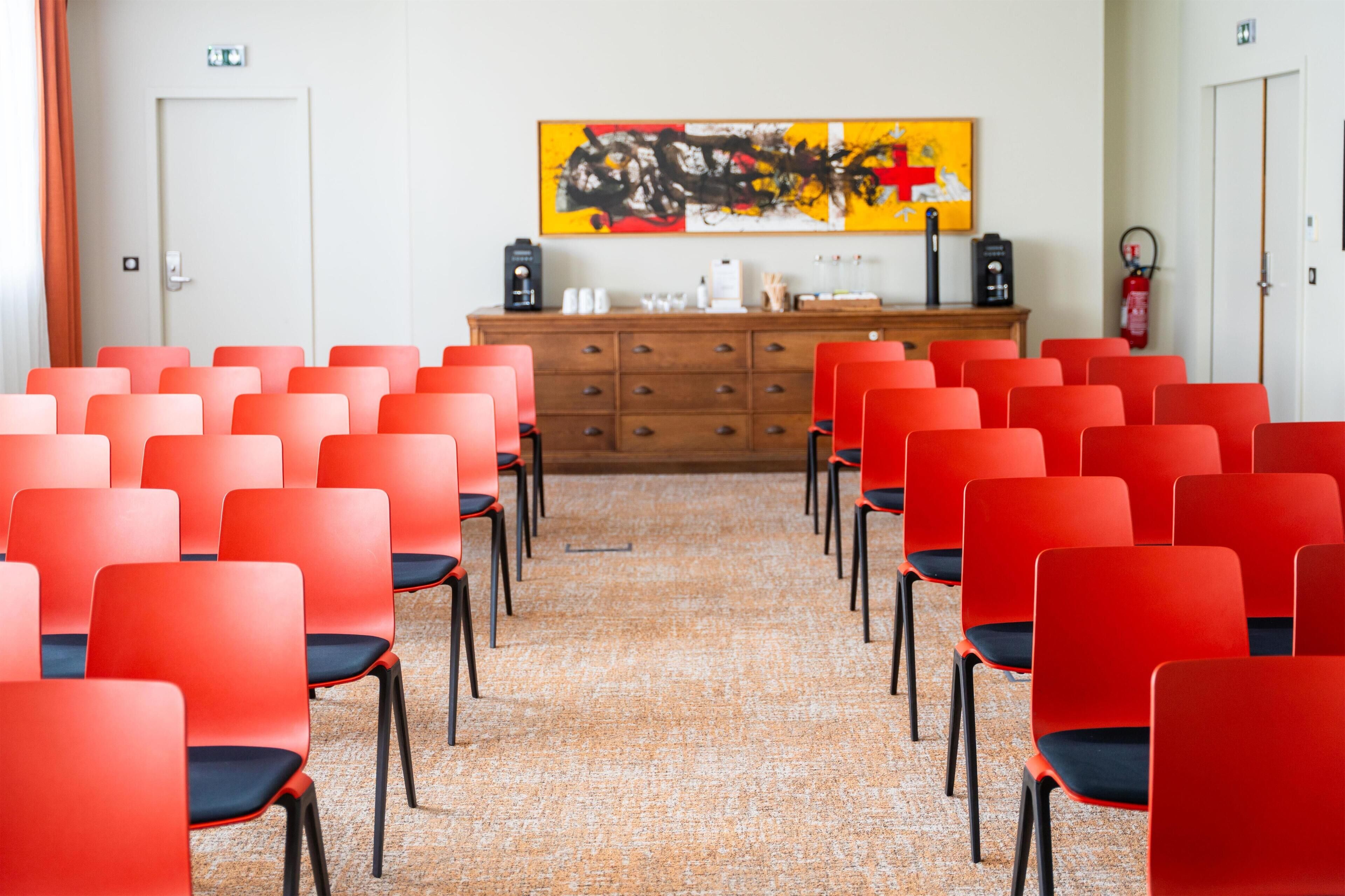 A conference room with rows of red chairs, a wooden cabinet with items on top, and a large abstract painting on the wall.