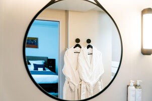 Un espejo redondo que refleja una habitación con dos batas blancas colgadas en una pared, una cama y un cuadro.