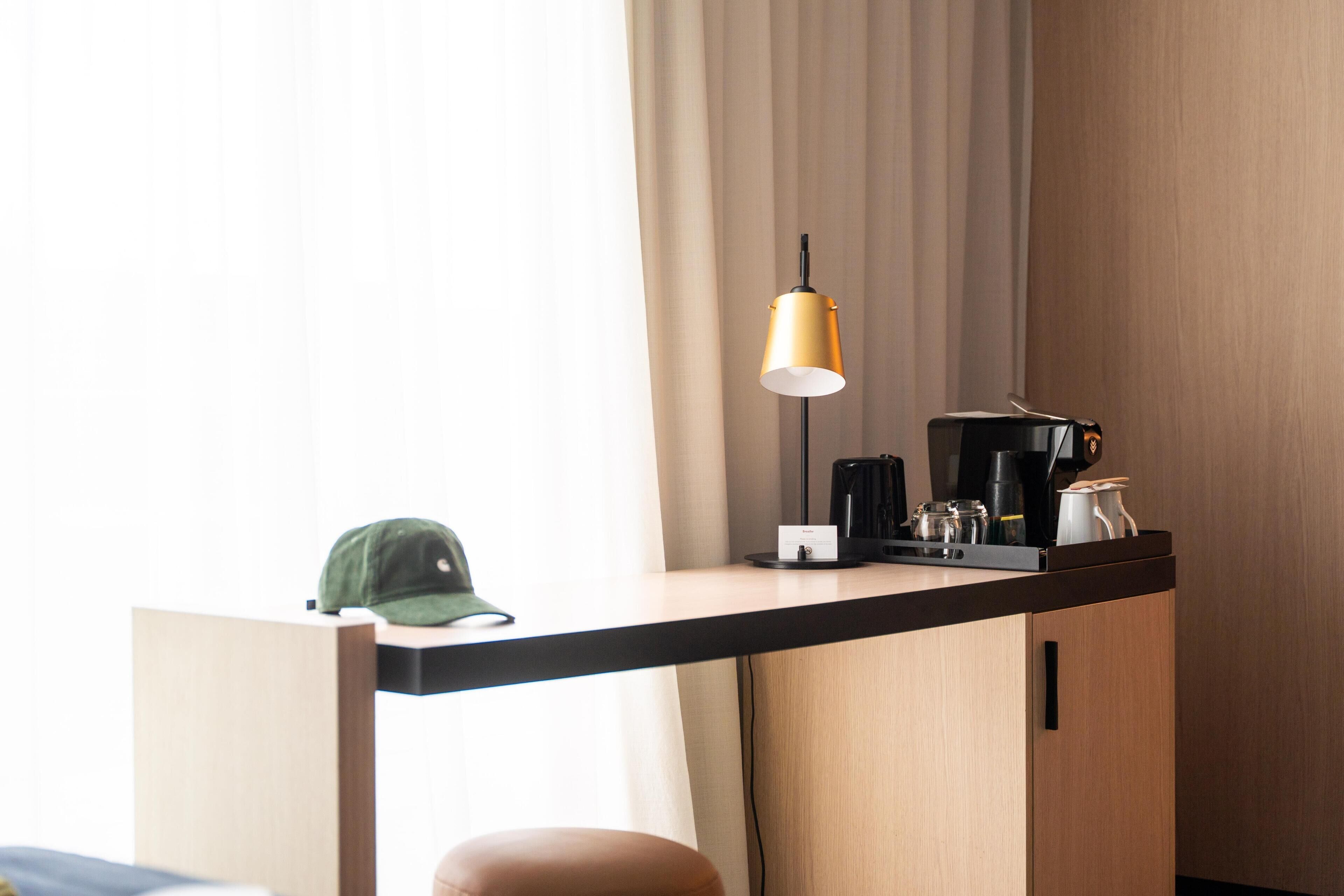A hotel room with a desk, a lamp, a coffee maker, and a cap on the desk.