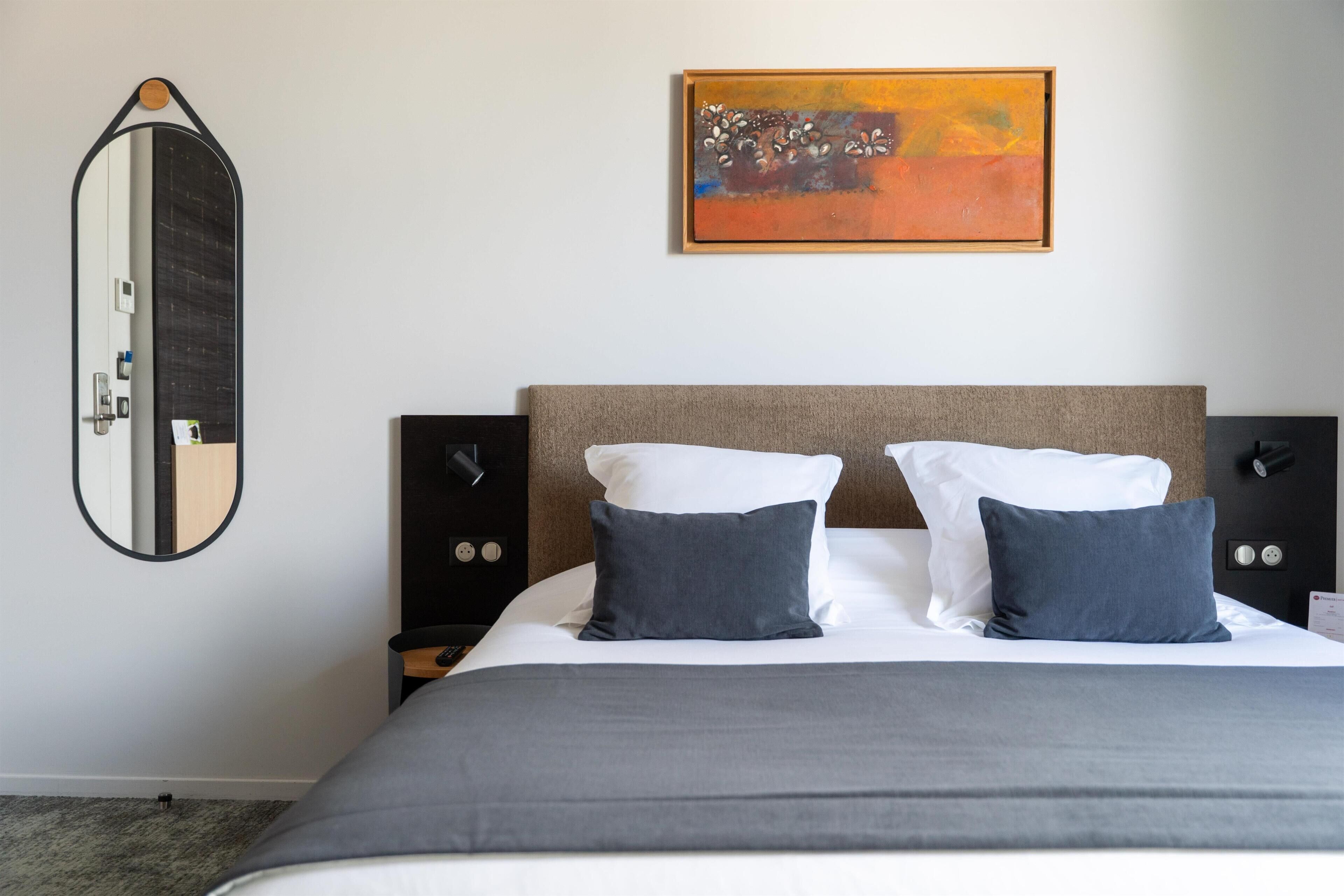 A hotel room with a bed, a bedside table, a wall-mounted mirror, and a painting.