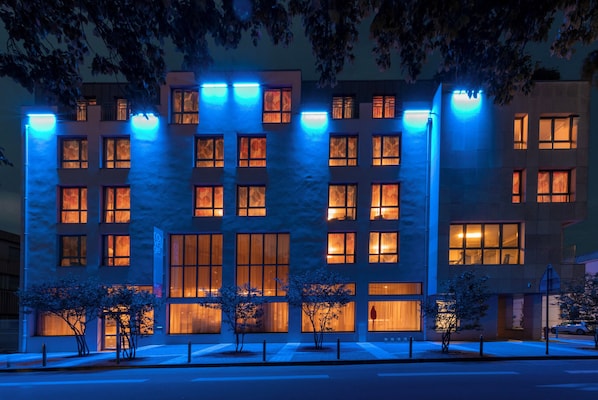 Un bâtiment moderne doté de nombreuses fenêtres et d’une façade plate, illuminé par un éclairage intérieur bleu et chaleureux.