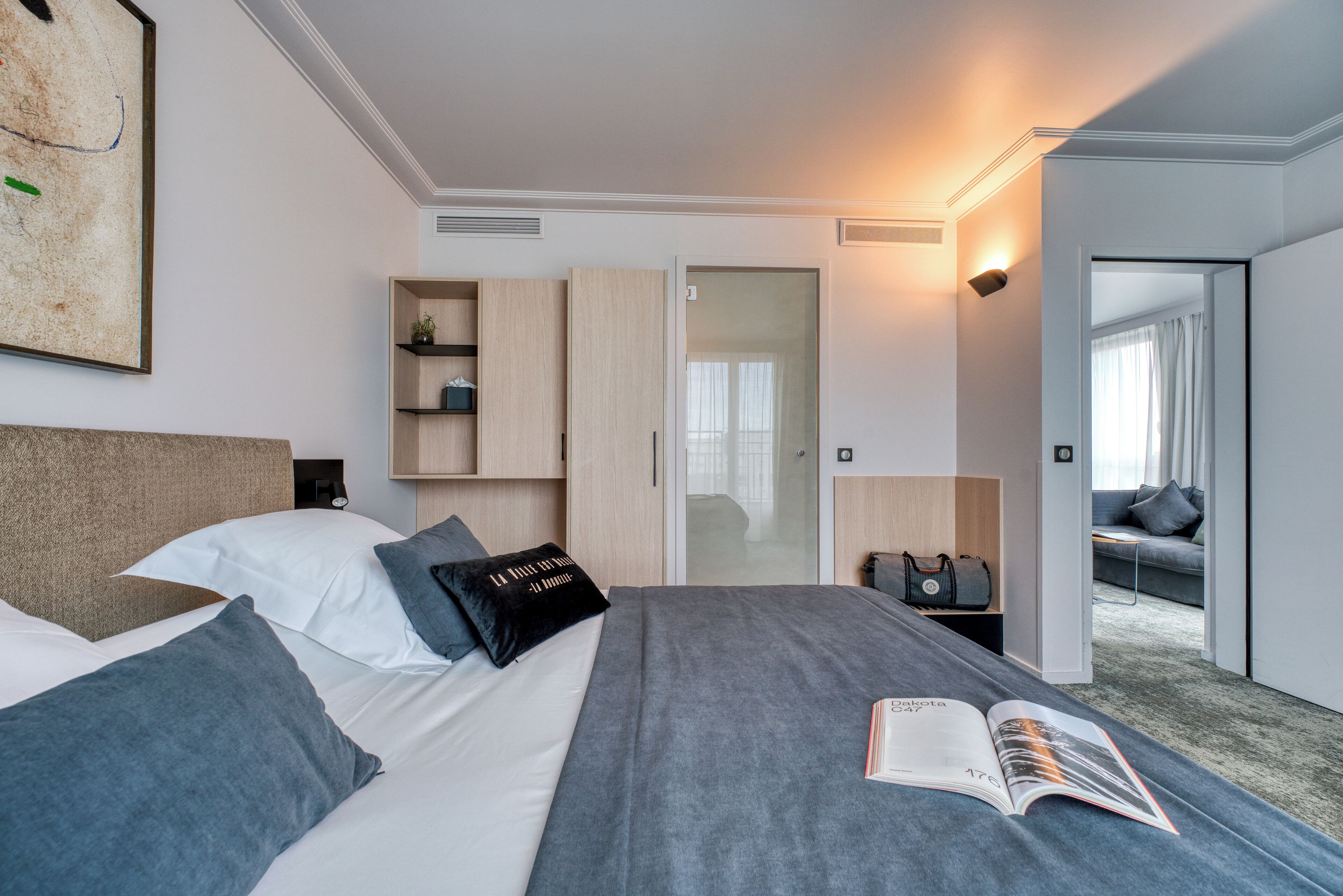 A modern hotel room with a large bed, a bedside table, a reading lamp, a wardrobe, and a view of the adjacent room.