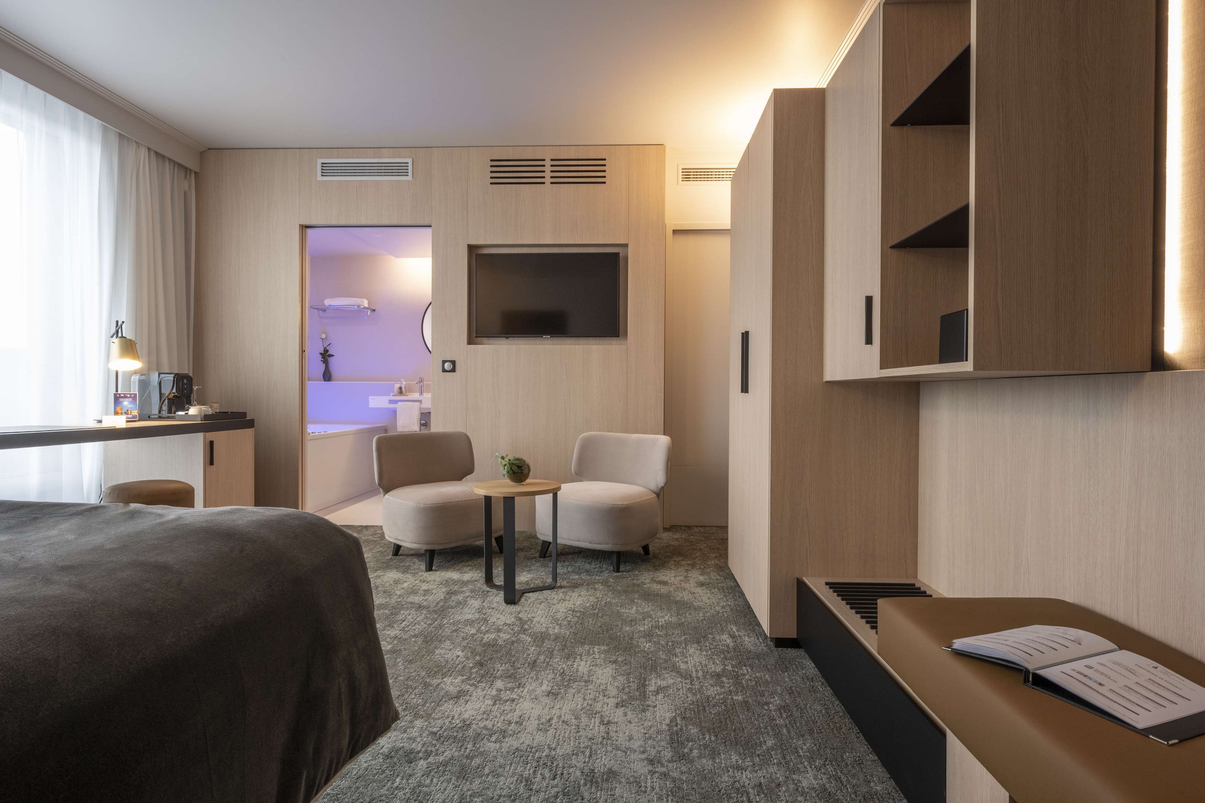 Habitación de hotel con cama, escritorio con lámpara, dos sillones, una mesita con planta, televisor montado en la pared y un piano.