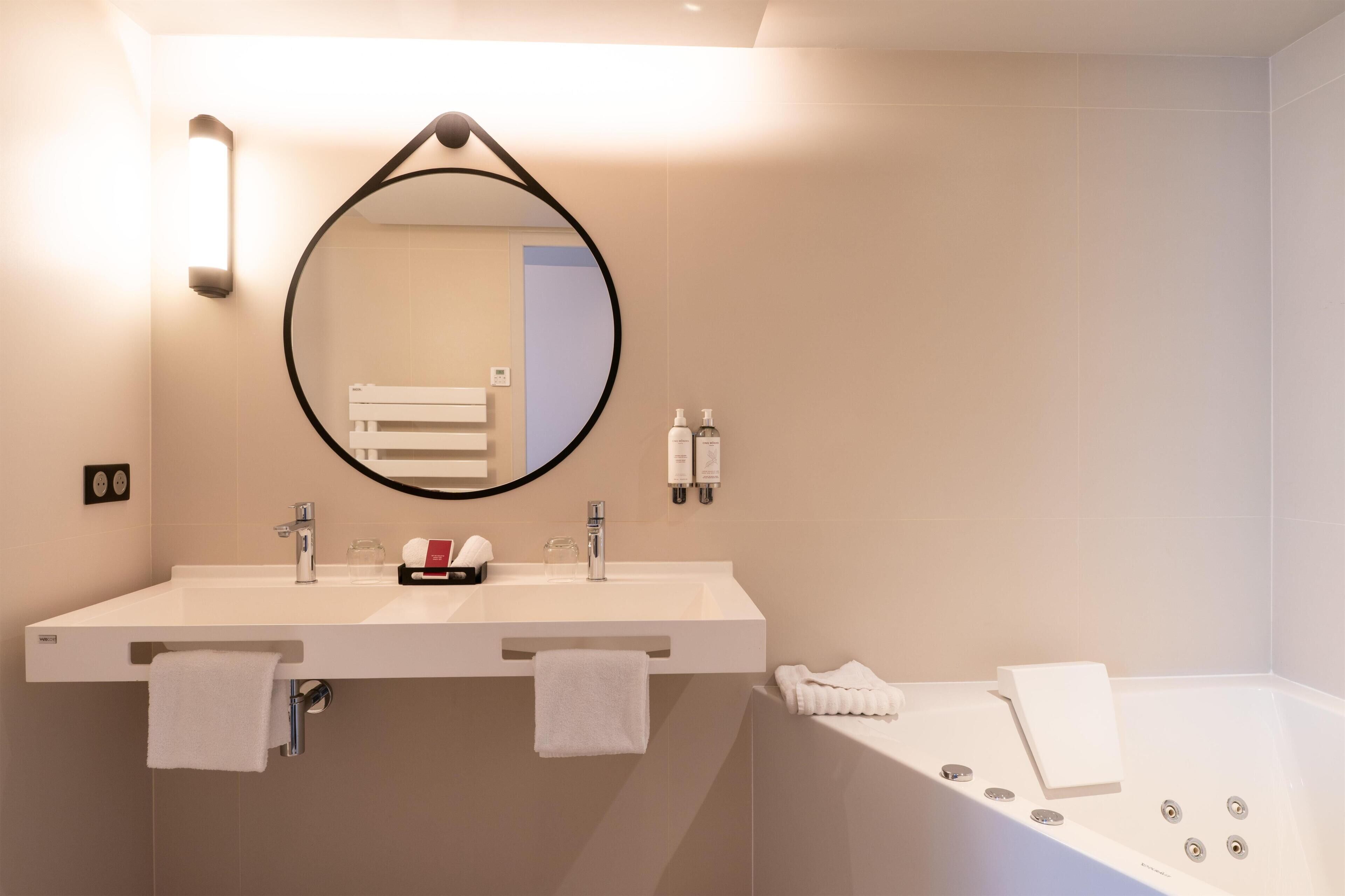 A modern bathroom with a double sink, a round mirror, and a bathtub.